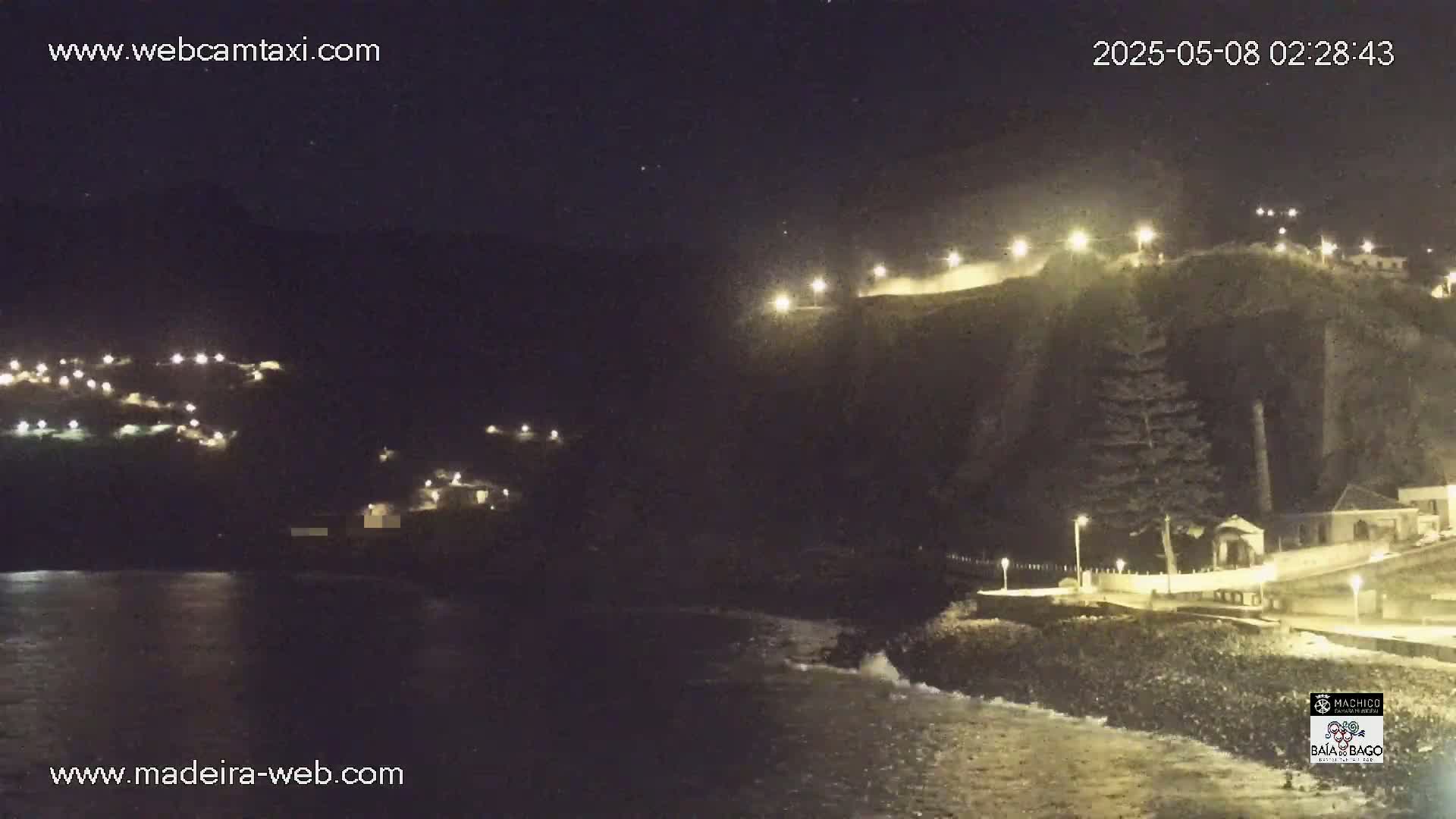 A nighttime coastal scene shows a dark body of water lapping against a rocky shore, with illuminated buildings and streets visible on a hillside above.
