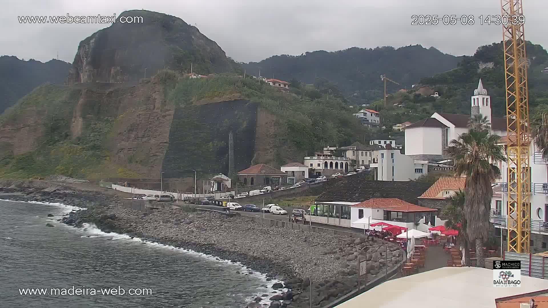 A rocky coastline with a town nestled against a steep, verdant cliff face under an overcast sky.
