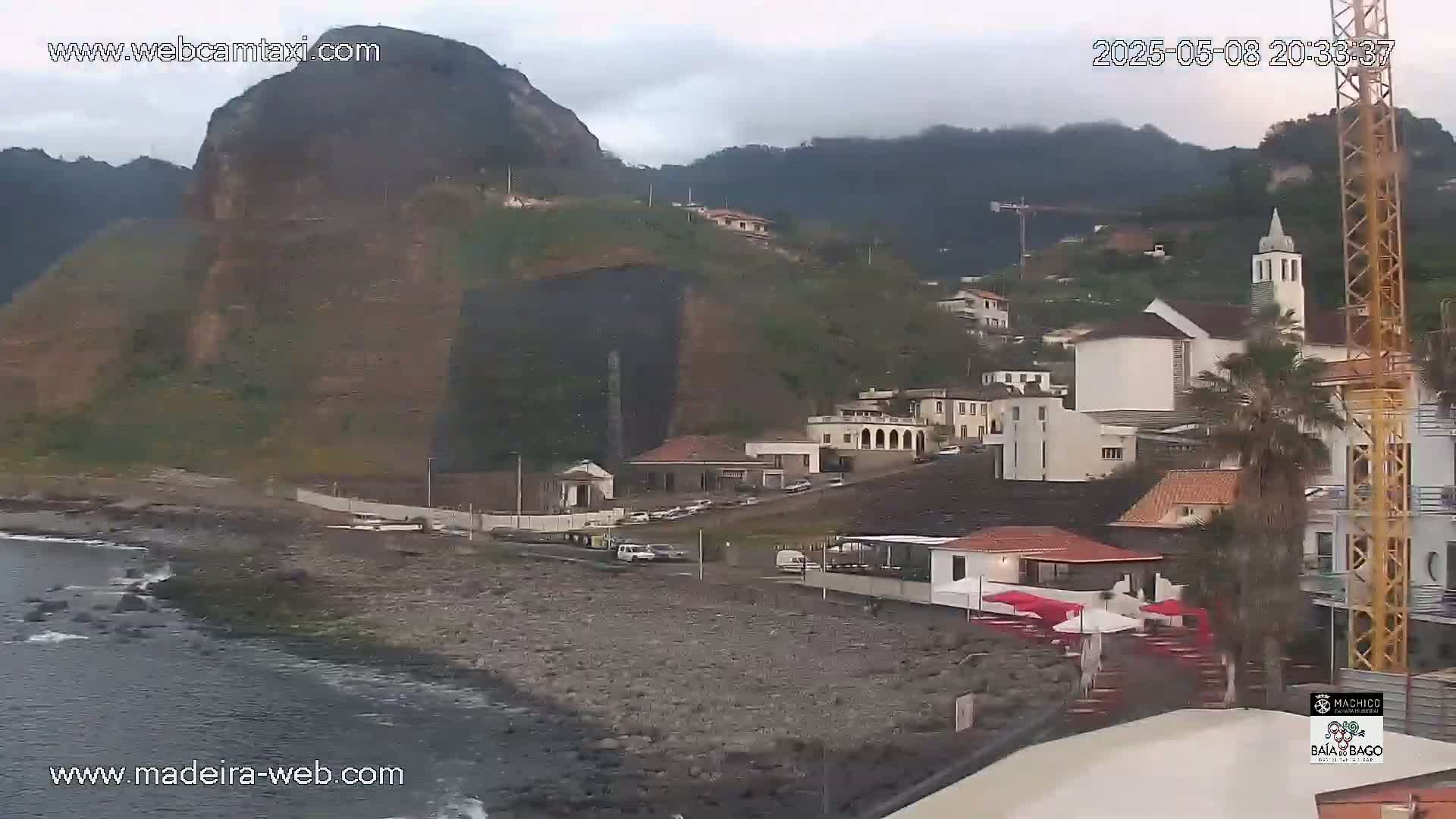 A rocky coastline with a village nestled against a steep, dark cliff and mountain range under an overcast sky.