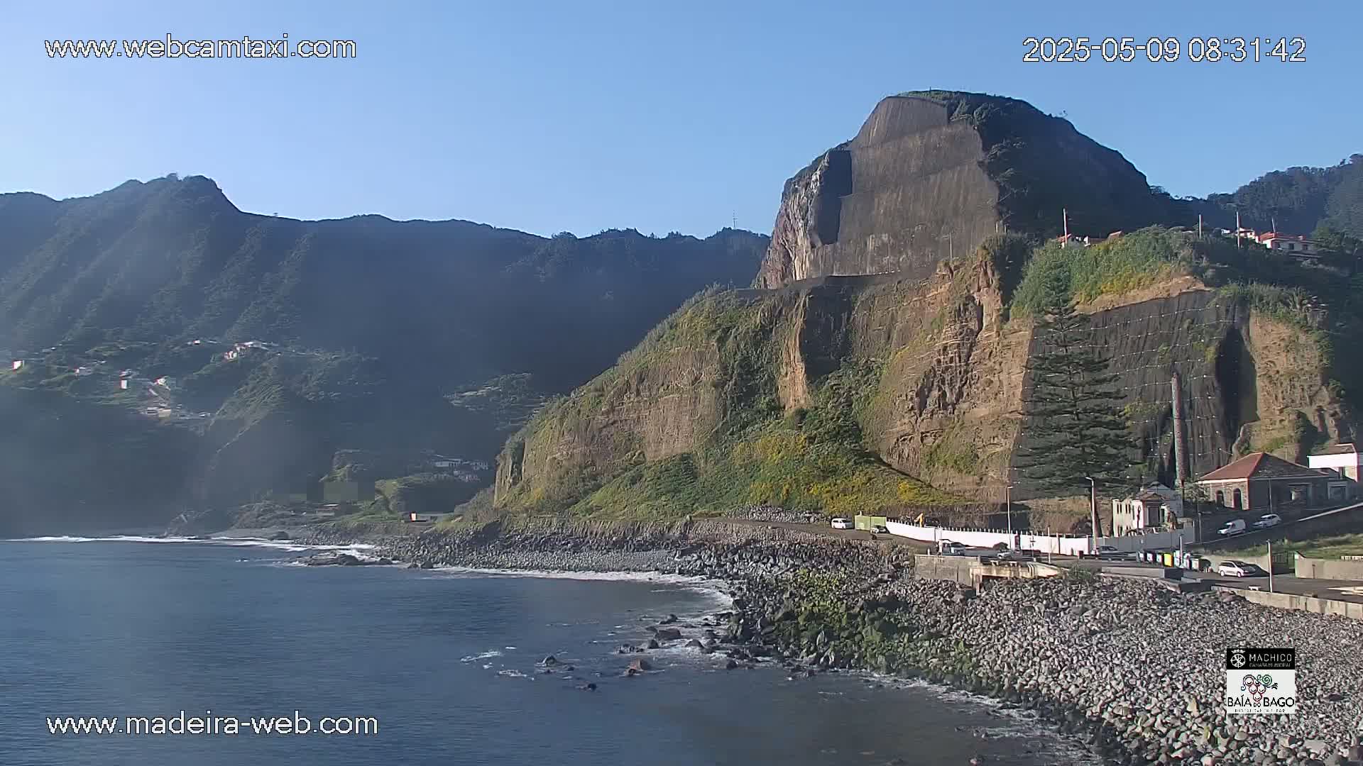 A rocky coastline with a steep cliff face, partially covered in netting, overlooks a calm ocean under a clear blue sky.