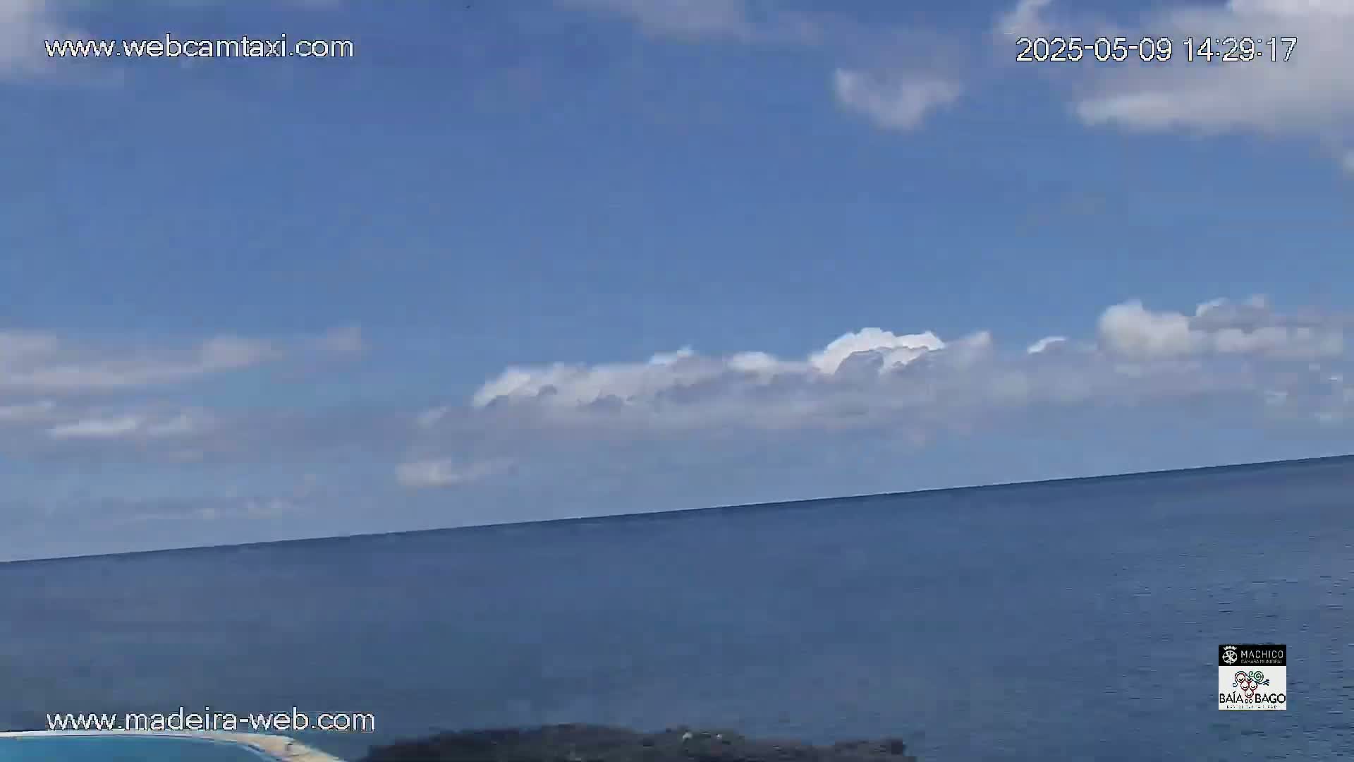 A mostly clear blue sky with some scattered clouds hangs above a calm, dark blue ocean.