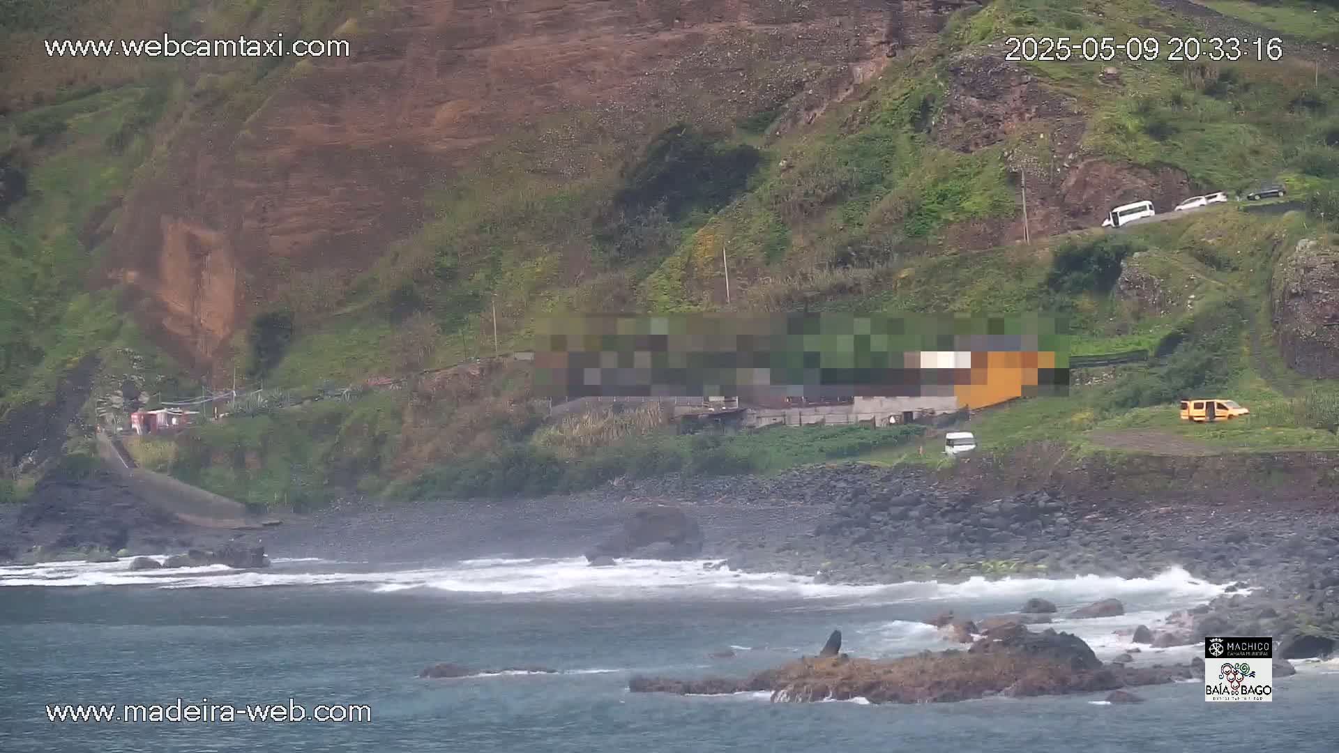A rocky coastline with a few buildings and vehicles is seen under overcast skies, with waves gently lapping at the shore.