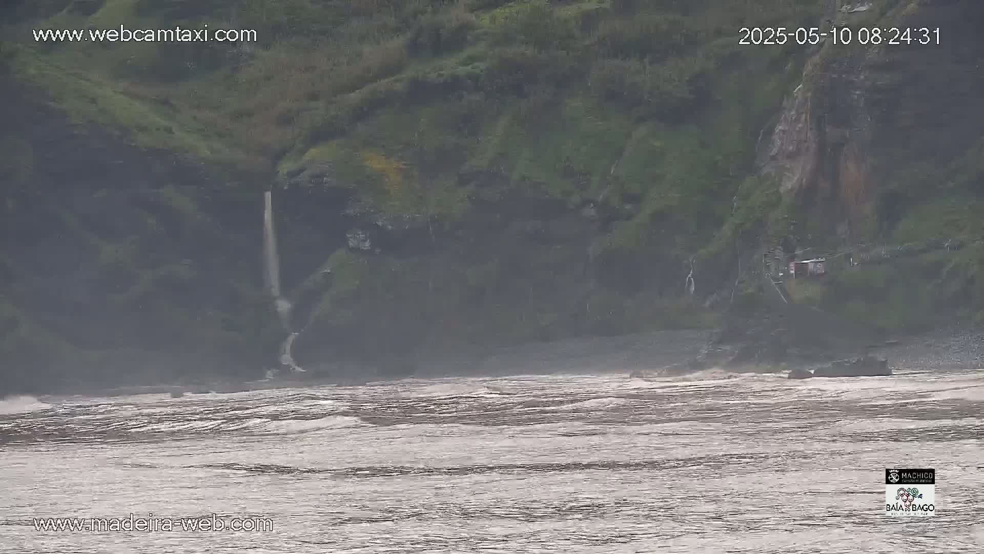 A waterfall cascades down a cliffside into a turbulent, grey ocean under an overcast sky.