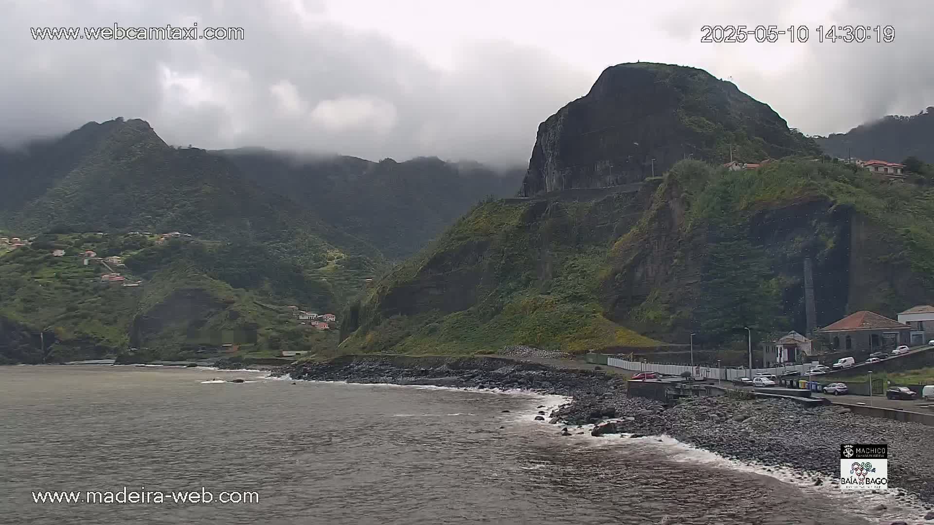 A rocky coastline with a village nestled into the green hills under a cloudy sky.