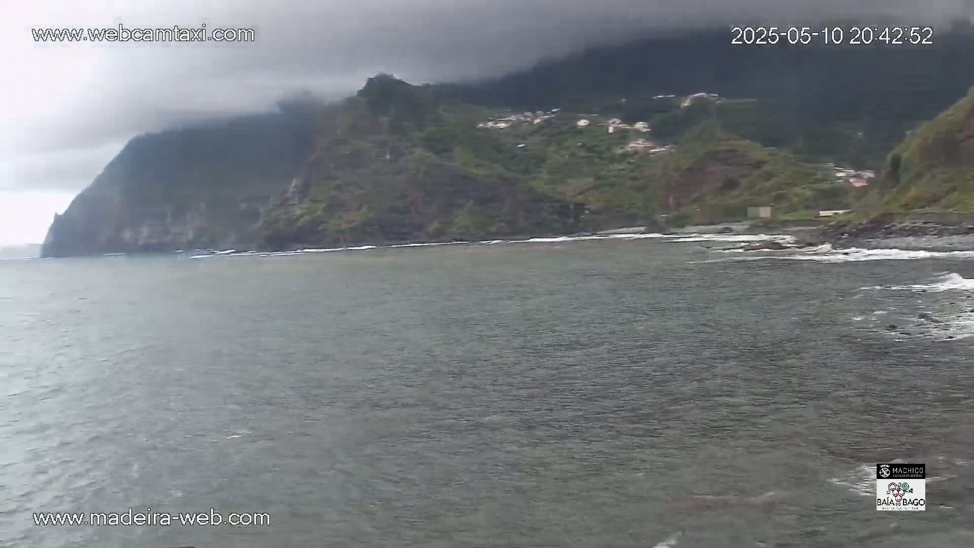 A dark, overcast sky hangs over a calm ocean that laps against a rocky shore lined with green hills and a small village.