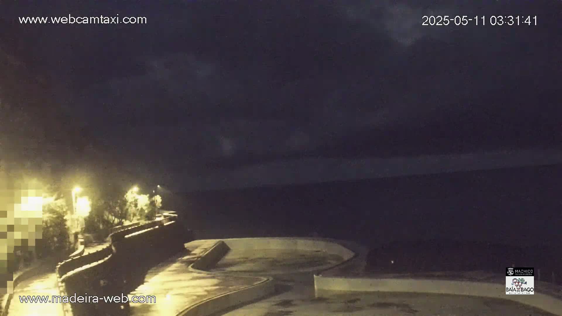A nighttime view of a paved waterfront area with some vegetation and streetlights, under a partly cloudy sky.
