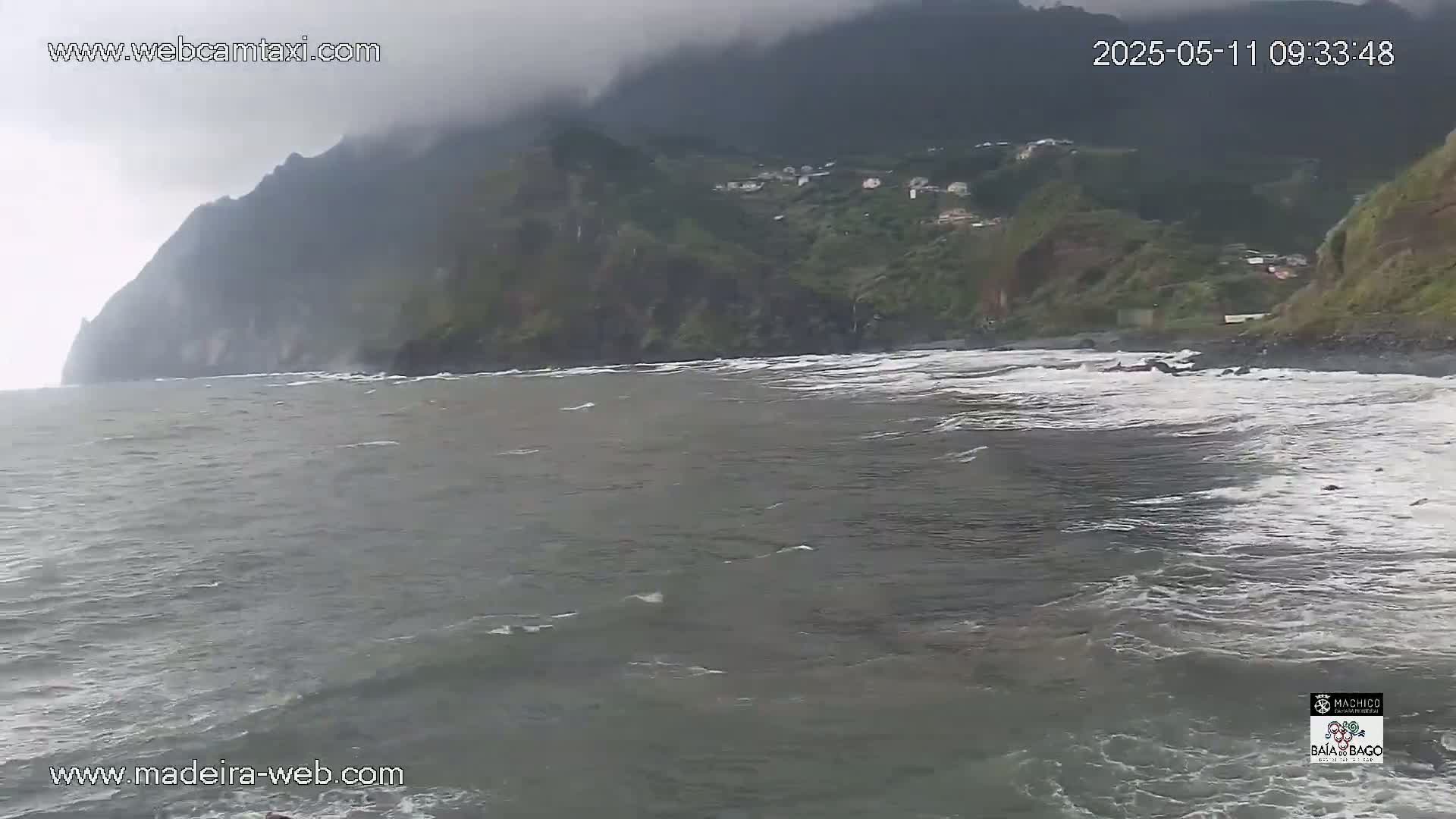 A dark, overcast day shows a rocky coastline with small waves lapping a dark-sand beach, and a small village nestled into the hillside in the distance.