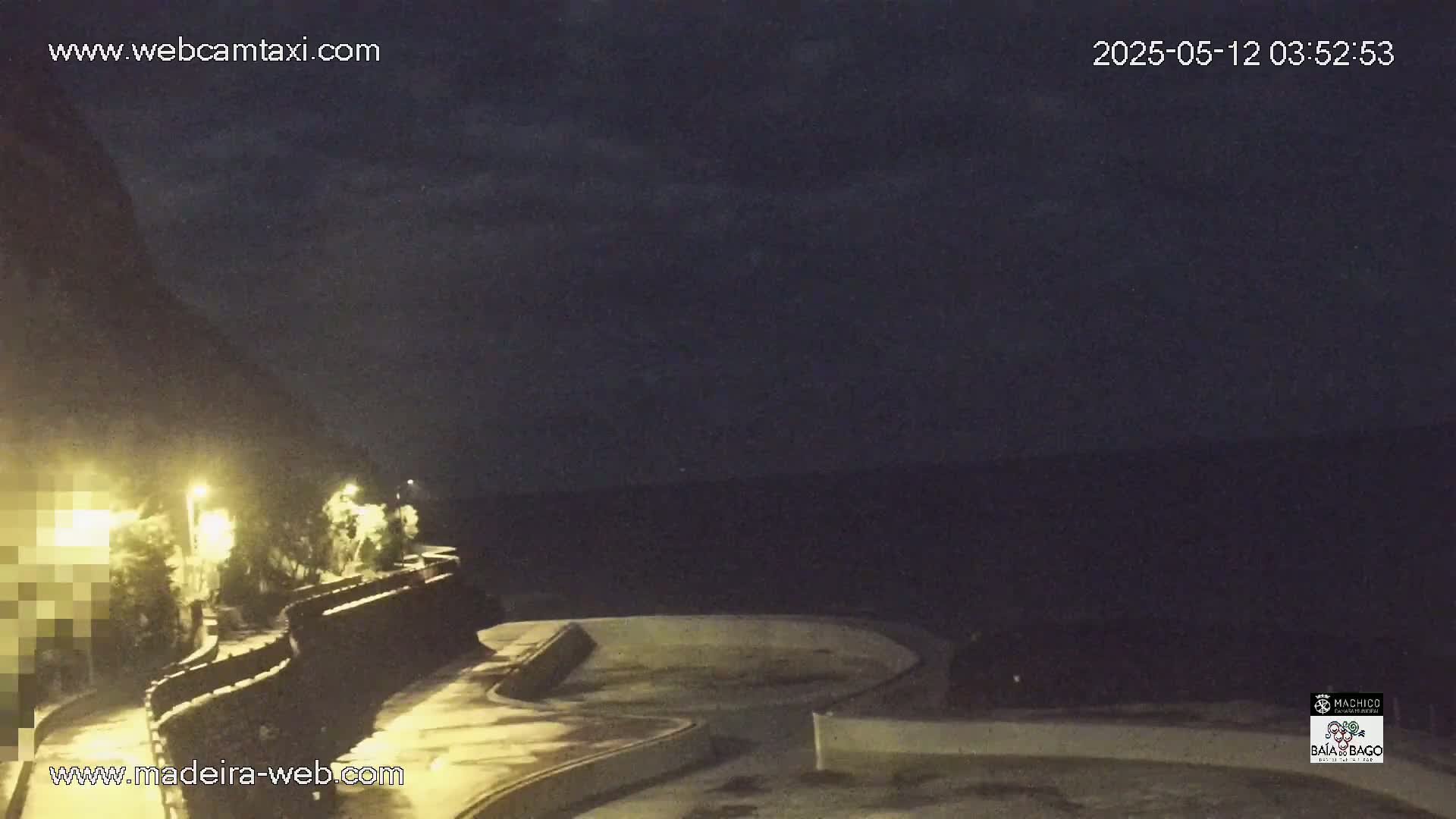 A nighttime coastal scene shows a dark ocean under a cloudy sky, with a curving roadway and stone wall illuminated by streetlights.