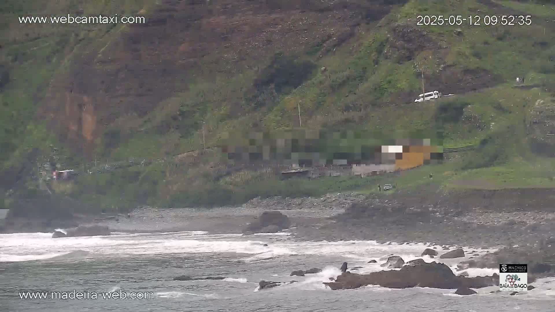 A rocky beach with small waves is visible, next to a hillside with some buildings and a road; the weather appears overcast.