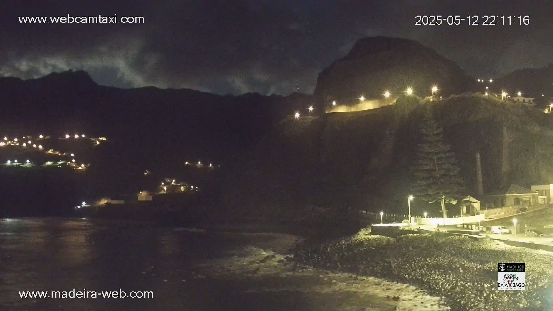 A nighttime coastal scene shows a dark, rocky cliff face illuminated by streetlights, with a small town nestled into the hillside and a dark body of water below.