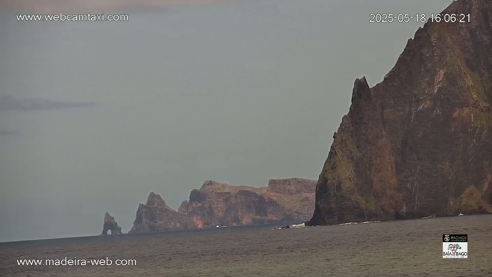 Dark, rocky cliffs rise from a calm, gray sea under an overcast sky.