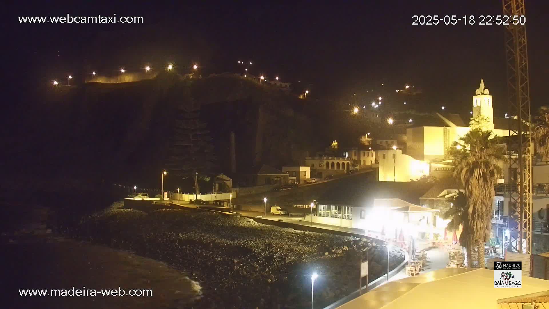 A nighttime coastal town scene shows buildings lit up along a rocky shoreline under a clear, dark sky.