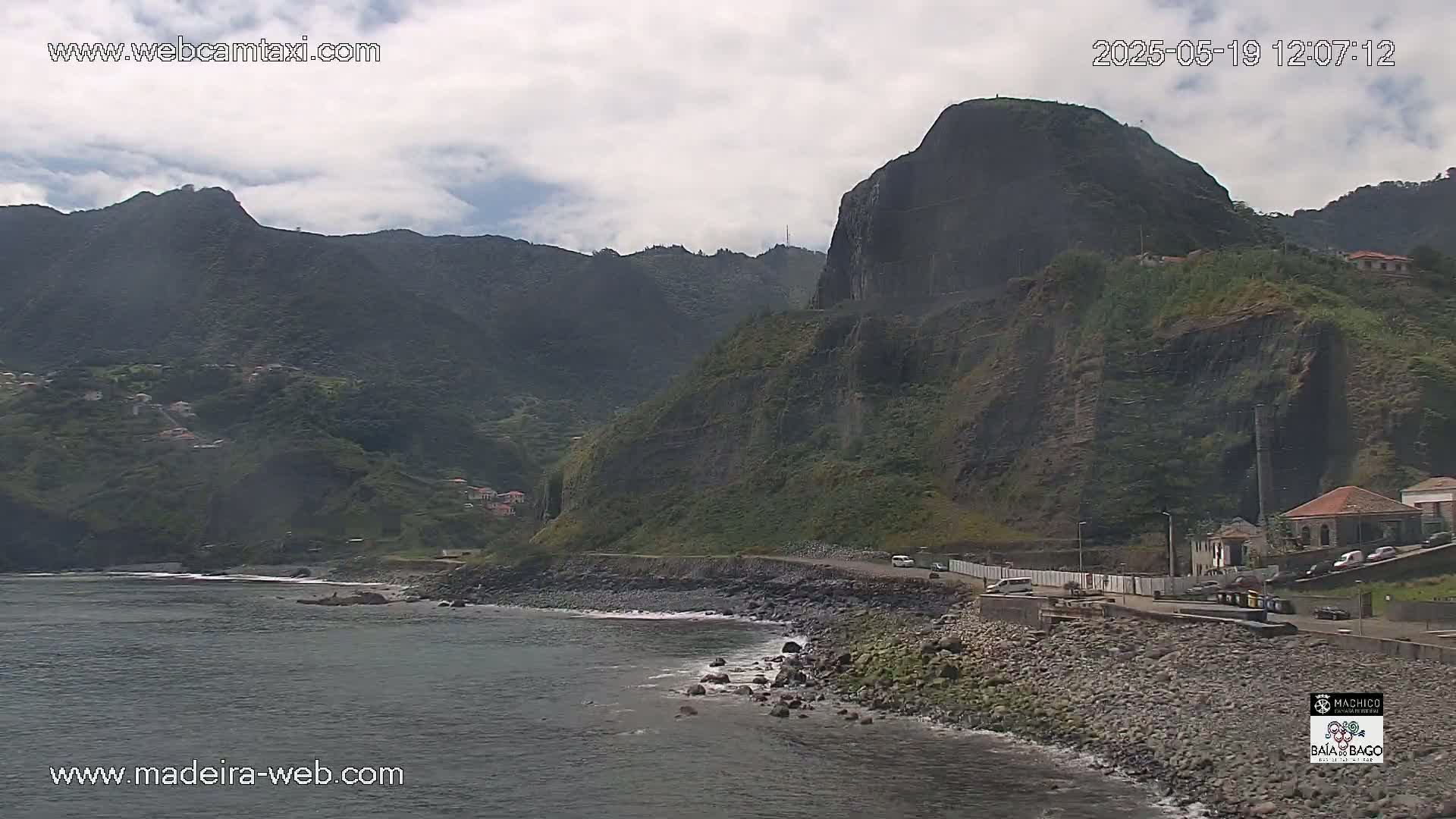 A rocky coastline with dark-colored cliffs and a few buildings is visible under a mostly cloudy sky.