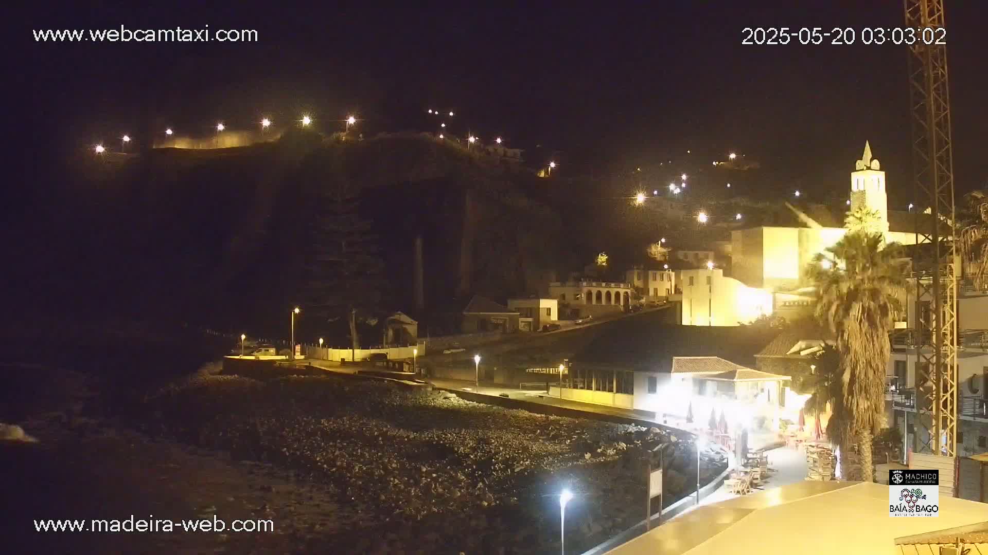 A nighttime view of a coastal town, illuminated by streetlights, with a rocky shoreline and buildings nestled against a hillside.