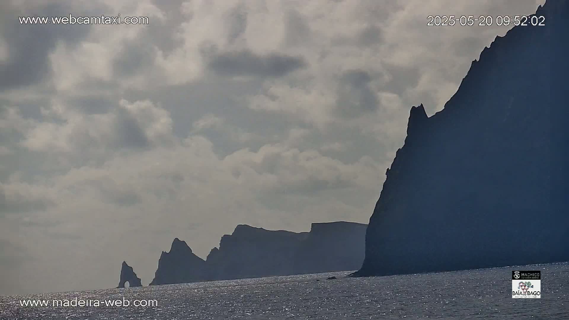 Dark, rocky cliffs rise from a calm, ocean under a cloudy sky.
