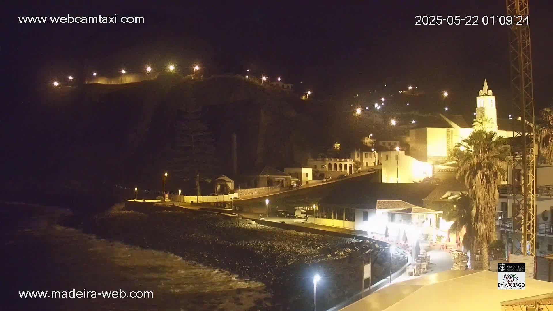 A nighttime coastal village scene shows a rocky beach with gentle waves, illuminated buildings on a hillside, and a prominent church tower.