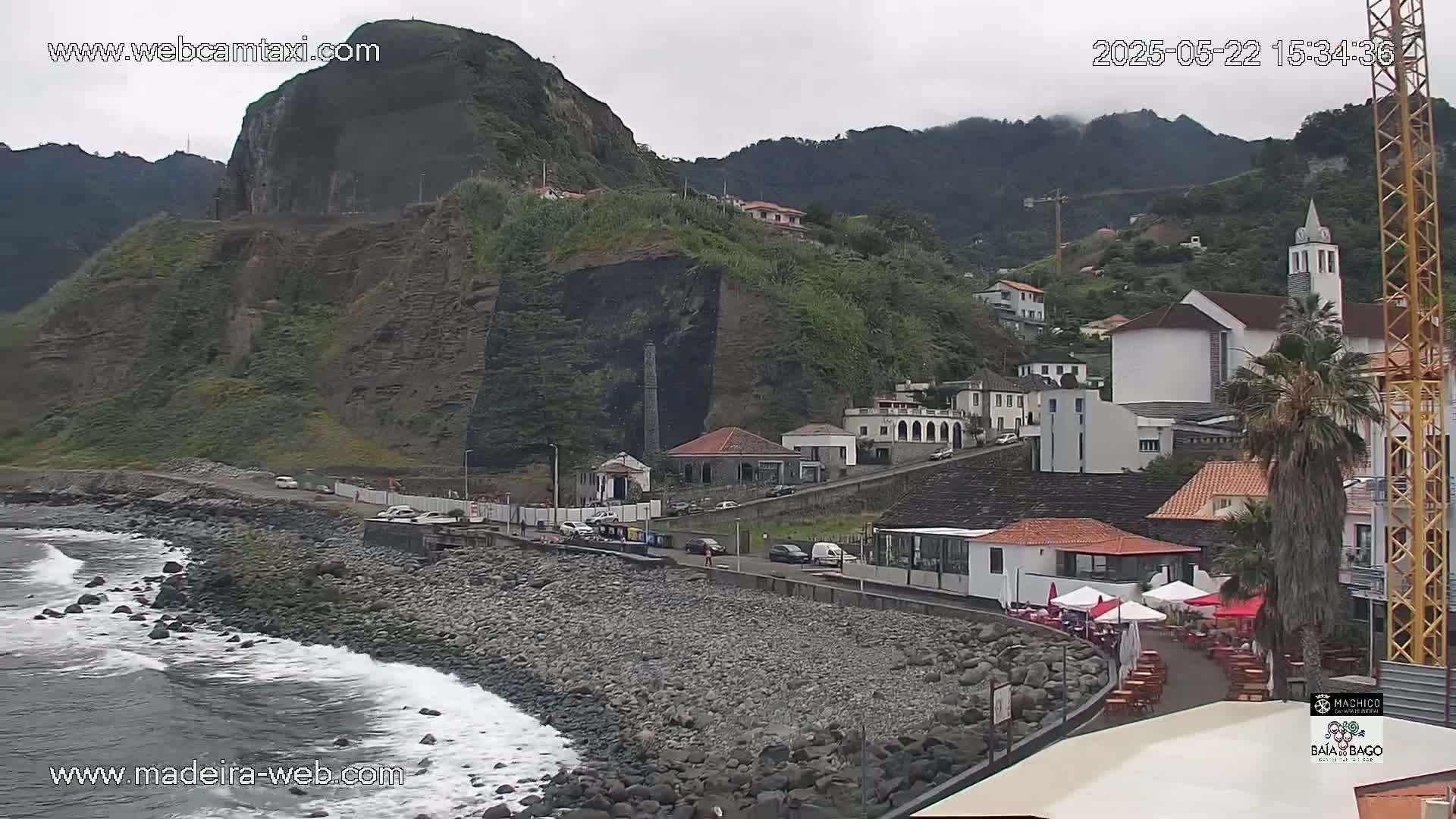 A rocky coastline with a small town nestled against a steep, green hillside under an overcast sky, with gentle waves lapping the shore.