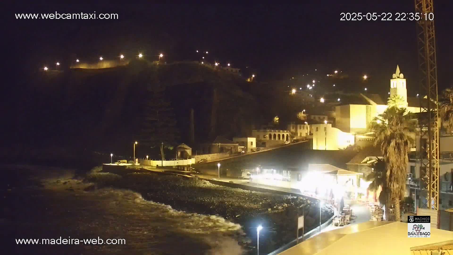 A nighttime coastal town, illuminated by streetlights, shows a rocky shore with waves gently lapping against it; buildings climb a hill behind the town's waterfront.