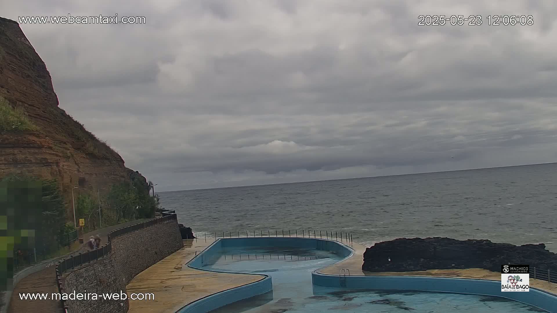 An outdoor view shows a coastal scene with a curving, light-blue swimming pool next to a dark-brown cliff, all under a mostly cloudy sky.