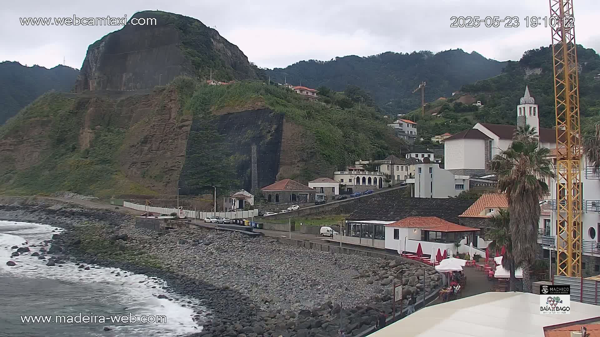 A rocky coastline with a village of white buildings nestled against a steep, green hillside under an overcast sky.
