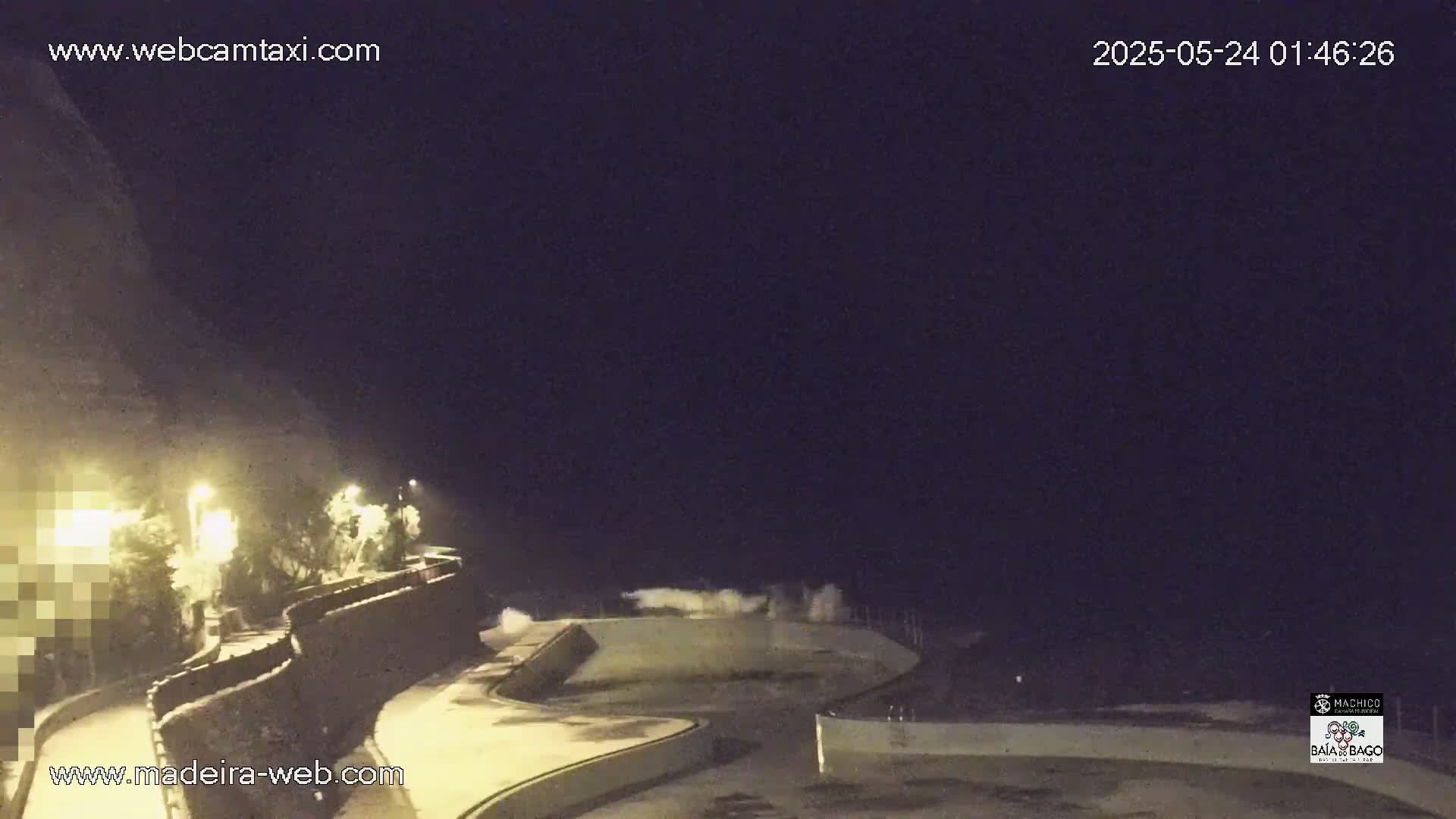 Large waves crash against a seawall at night, illuminating the area with white foam and splashing water.