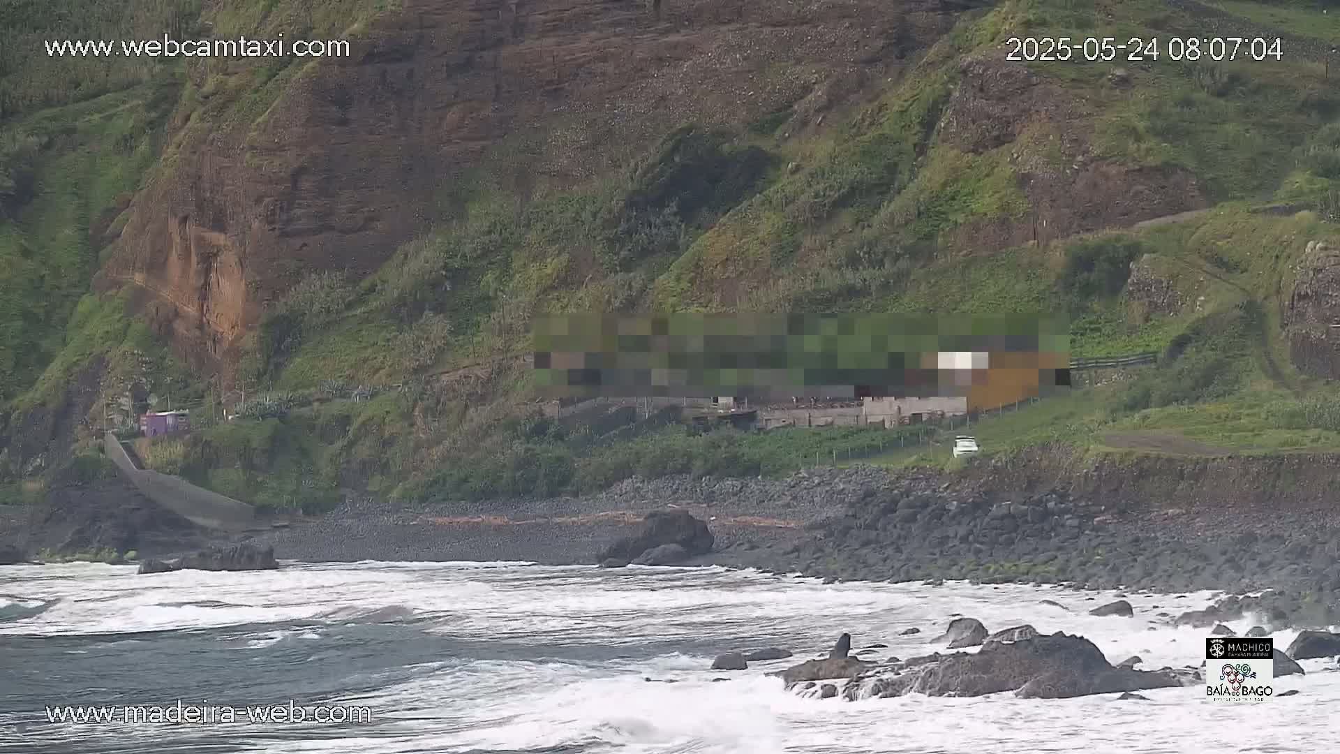 A rocky, dark-colored beach with moderate waves laps against the shore, beneath a green hillside with a building partially obscured by pixelation.