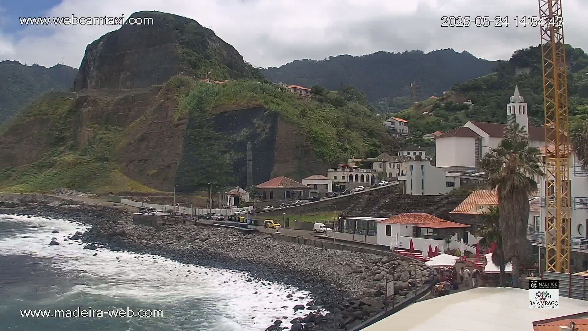 A rocky coastline with crashing waves features a village nestled against a steep, verdant hillside under a partly cloudy sky.
