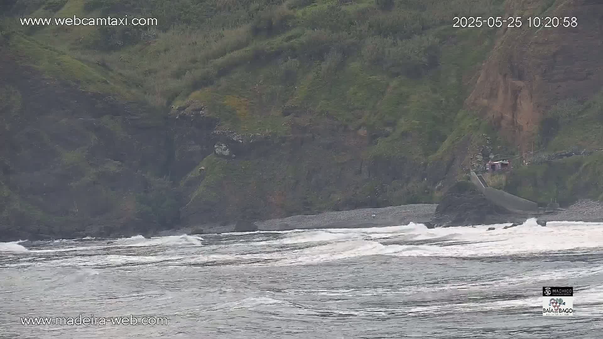 Waves crash on a rocky beach below a green, cliffside hillside on an overcast day.
