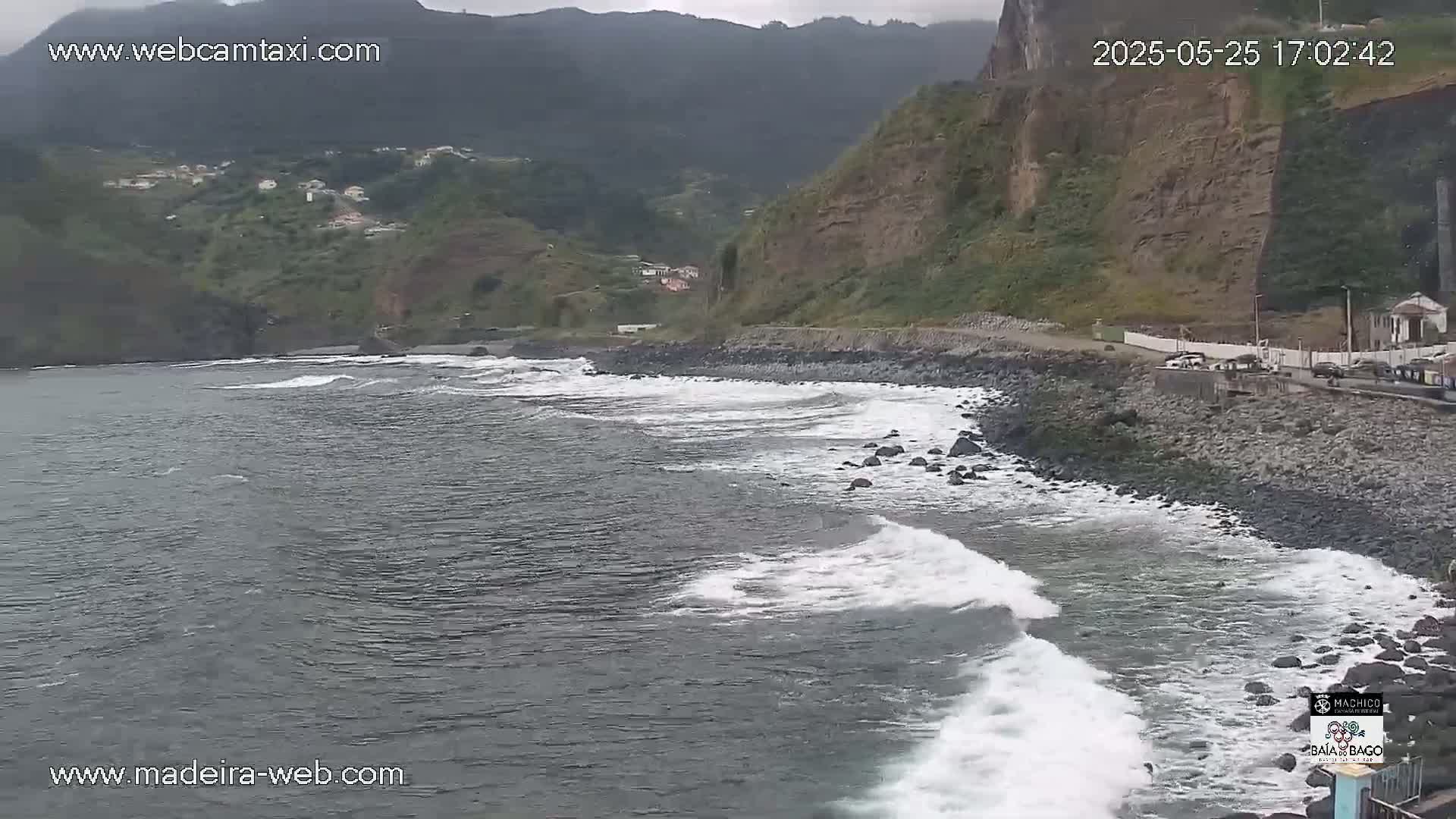 Waves crash on a rocky shore next to a road that runs along a steep, green hillside under an overcast sky.