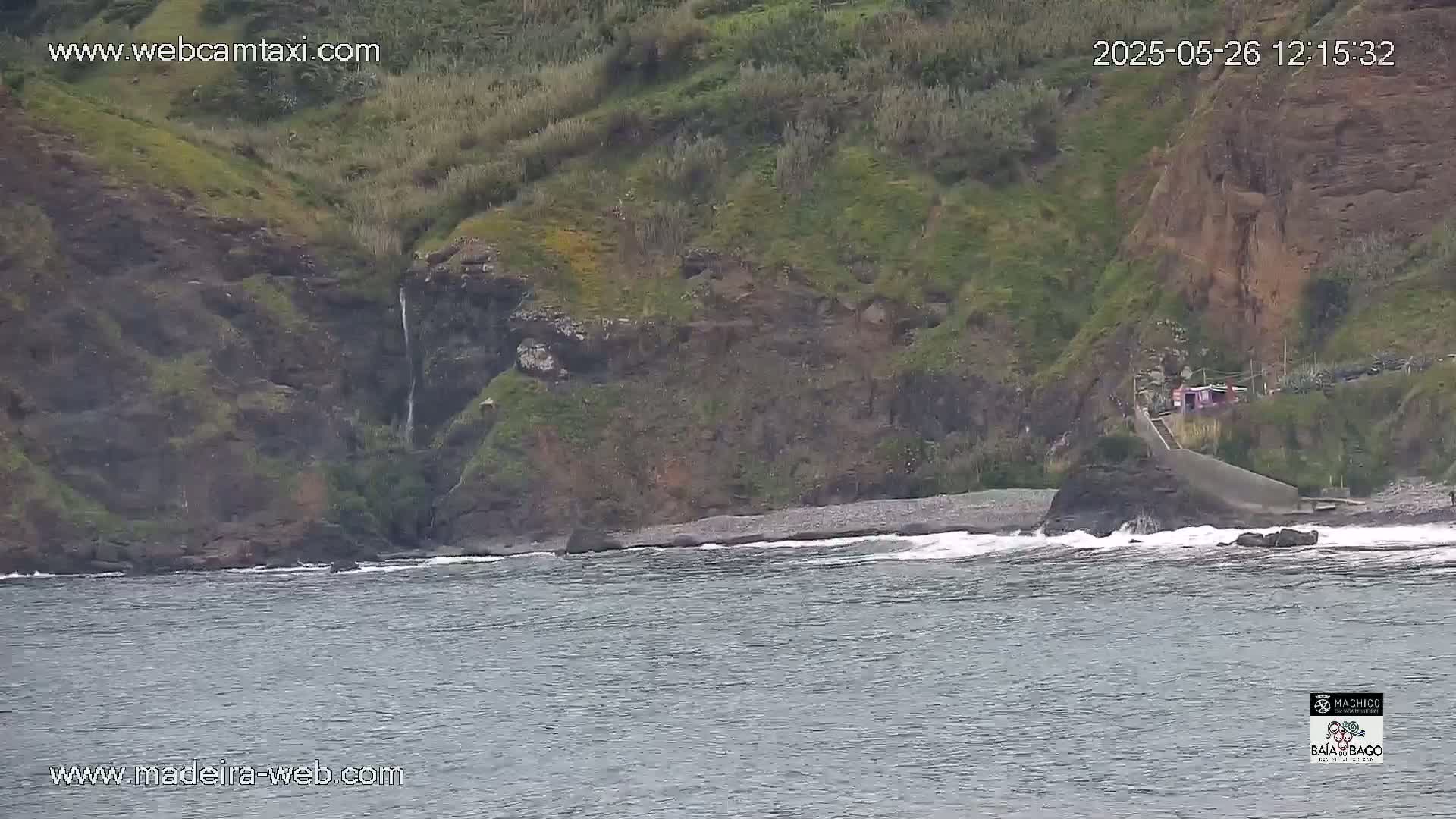 A rocky, green hillside meets a pebble beach and dark-grey ocean with small waves, featuring a small waterfall cascading down the cliff.