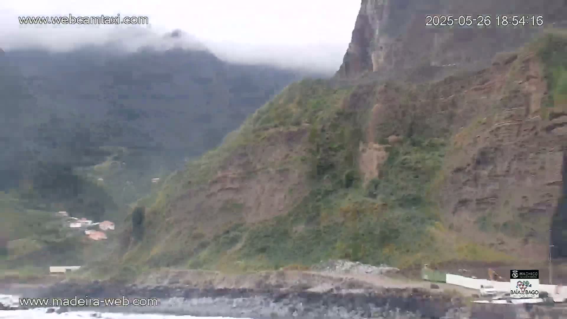 A misty, overcast view of a steep, rocky coastline with sparse vegetation, a small village nestled into the hillside, and a dark-colored beach at the base.