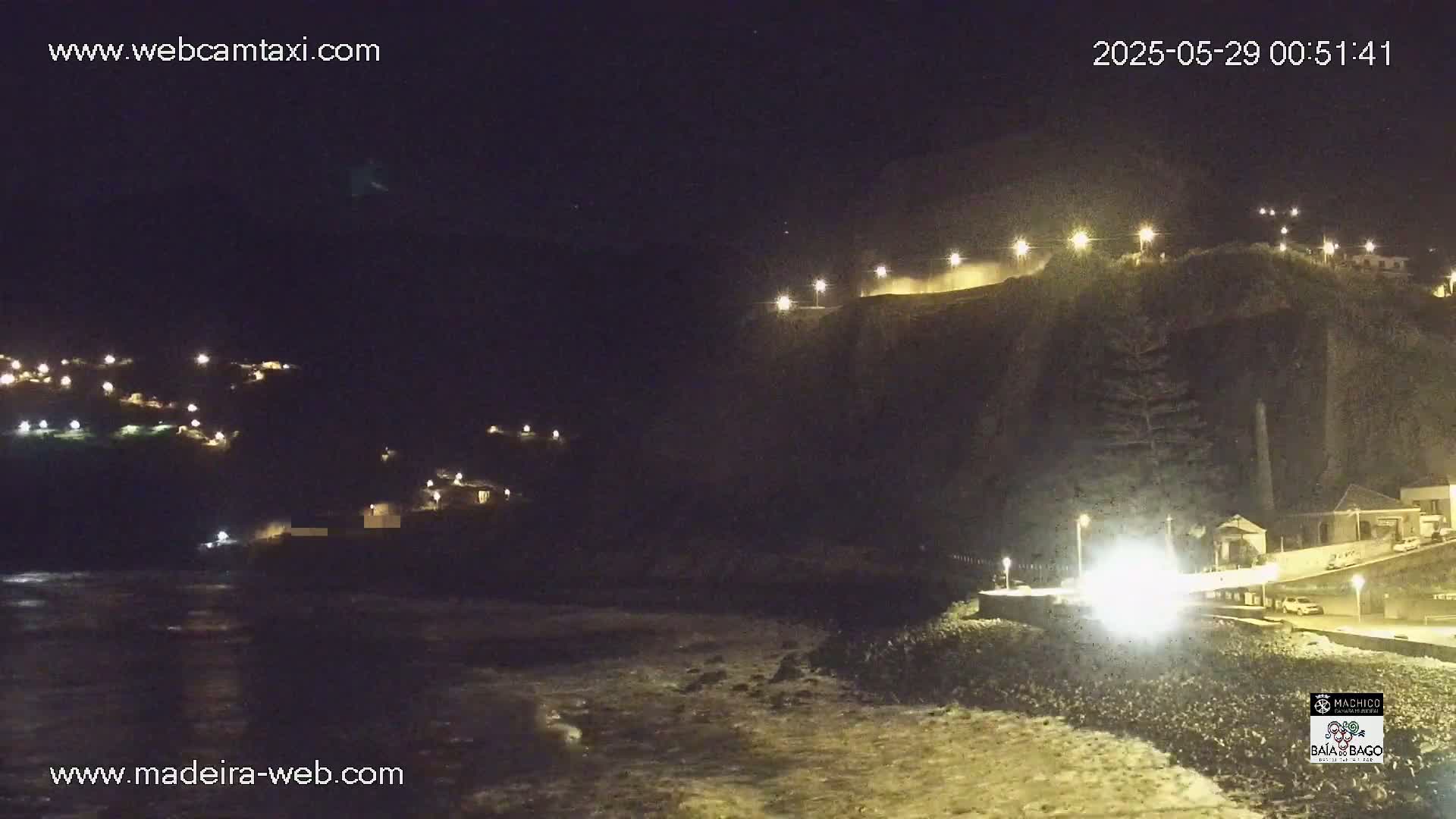 A nighttime coastal scene shows a dark cliff with lights along the top, a village nestled into the hillside, and waves breaking on the shore below.