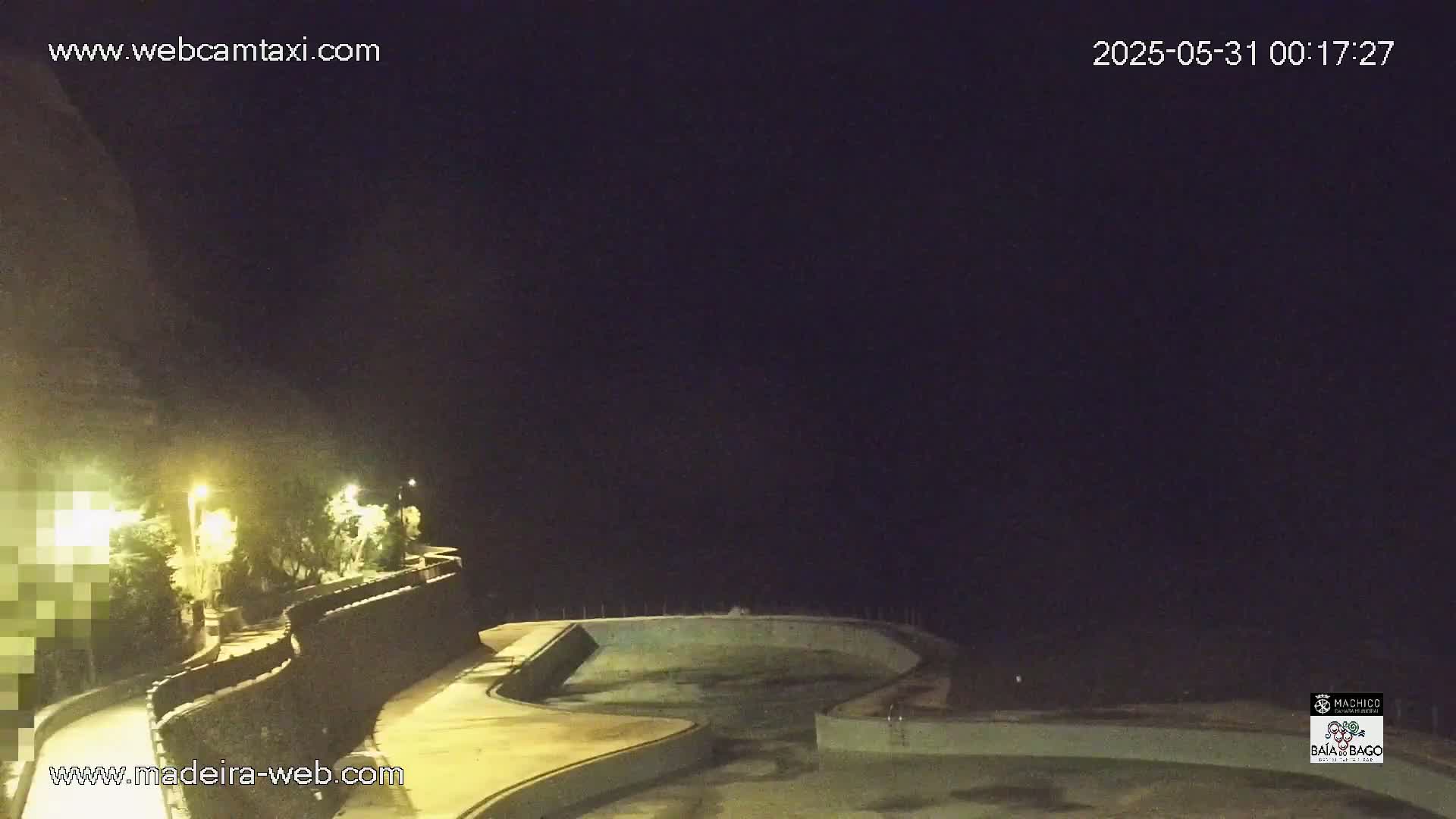 An empty, illuminated pool is situated at night next to a stone wall and some vegetation under a dark sky.
