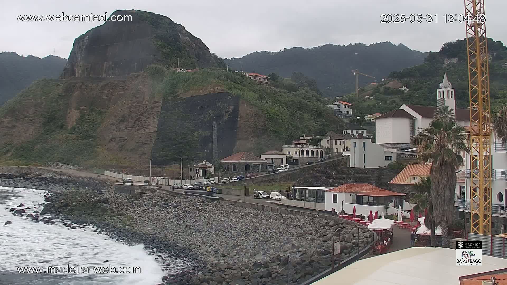 A rocky coastline with a small town nestled against a steep, green hillside under an overcast sky, with gentle waves lapping at the shore.