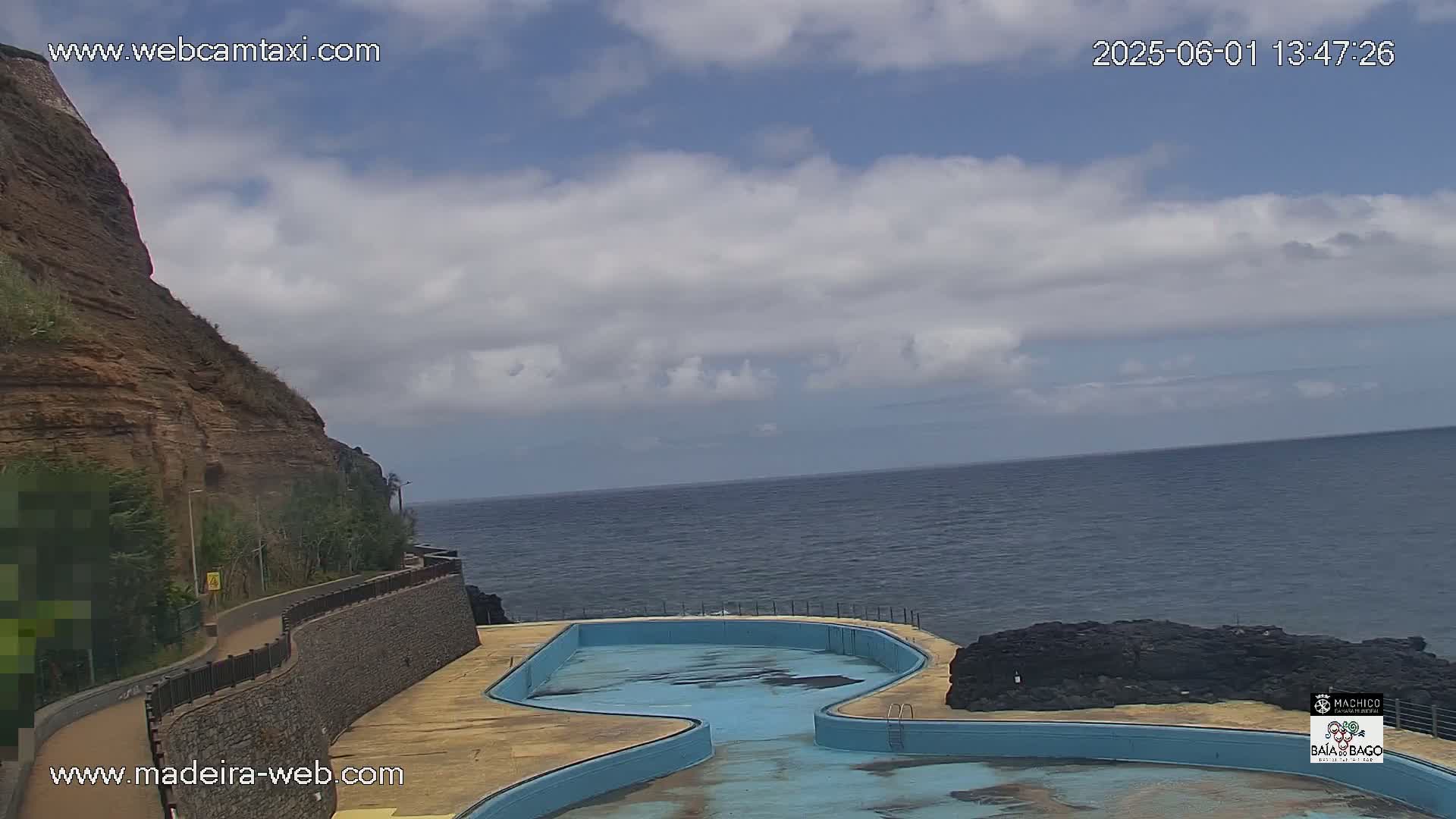 An outdoor scene shows a partially filled, light-blue pool next to a rocky cliff overlooking a calm ocean under a partly cloudy sky.
