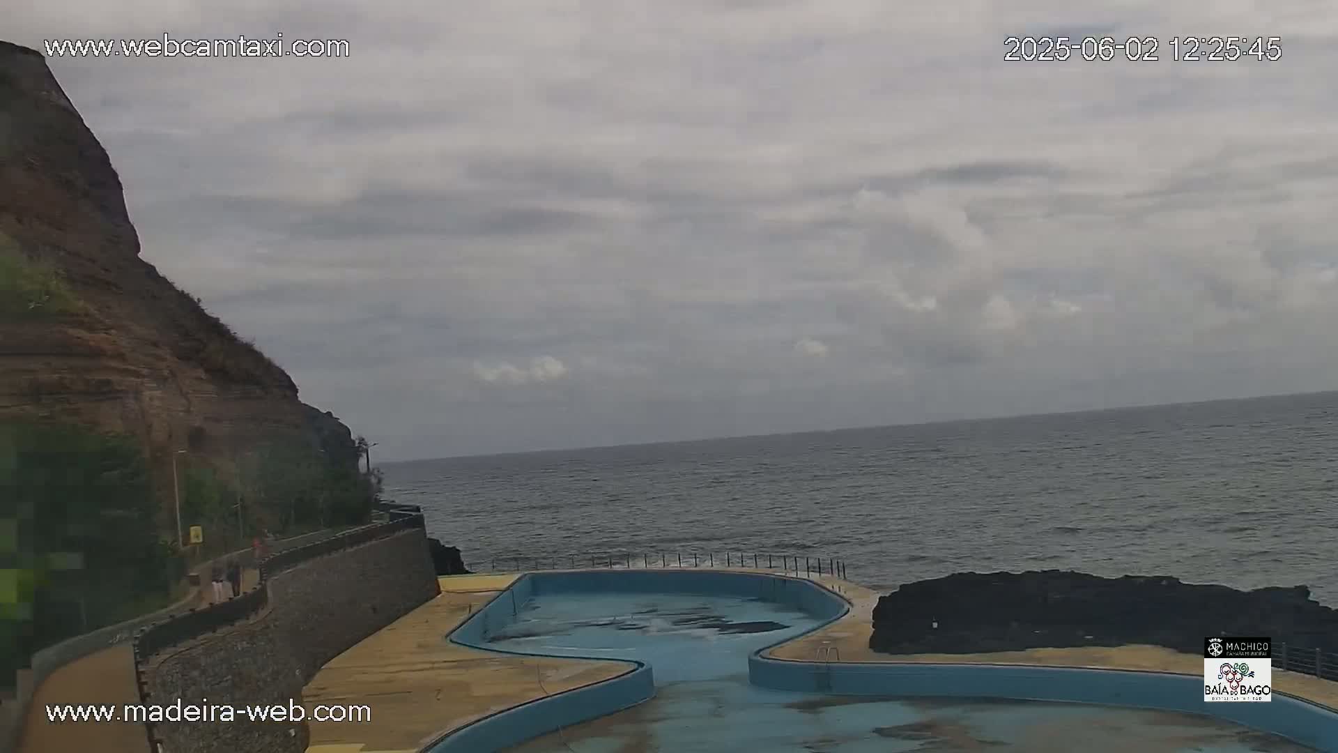 An outdoor scene shows a rocky cliff overlooking an ocean under a cloudy sky, with an empty, irregularly shaped pool situated between the cliff and the water.