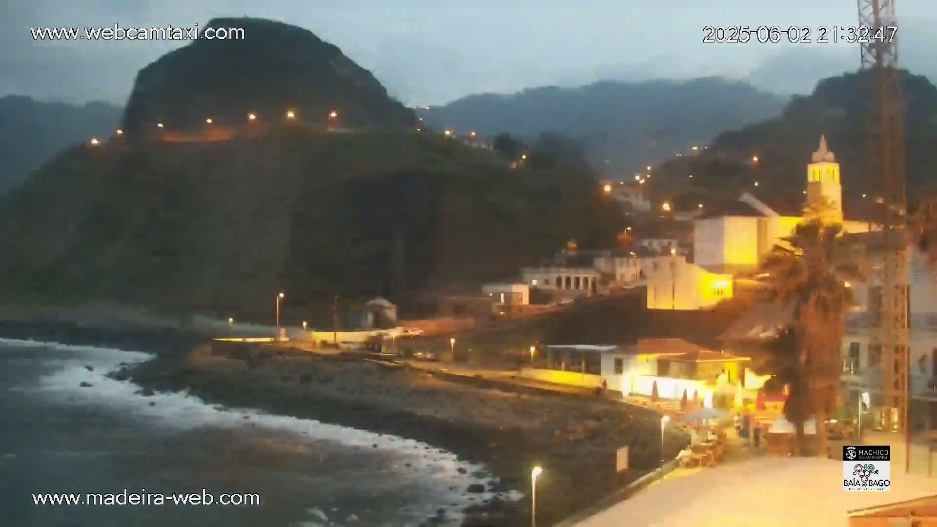 A coastal town at night, illuminated by streetlights, sits nestled against a dark, mountainous backdrop under a twilight sky with gentle waves lapping the shore.