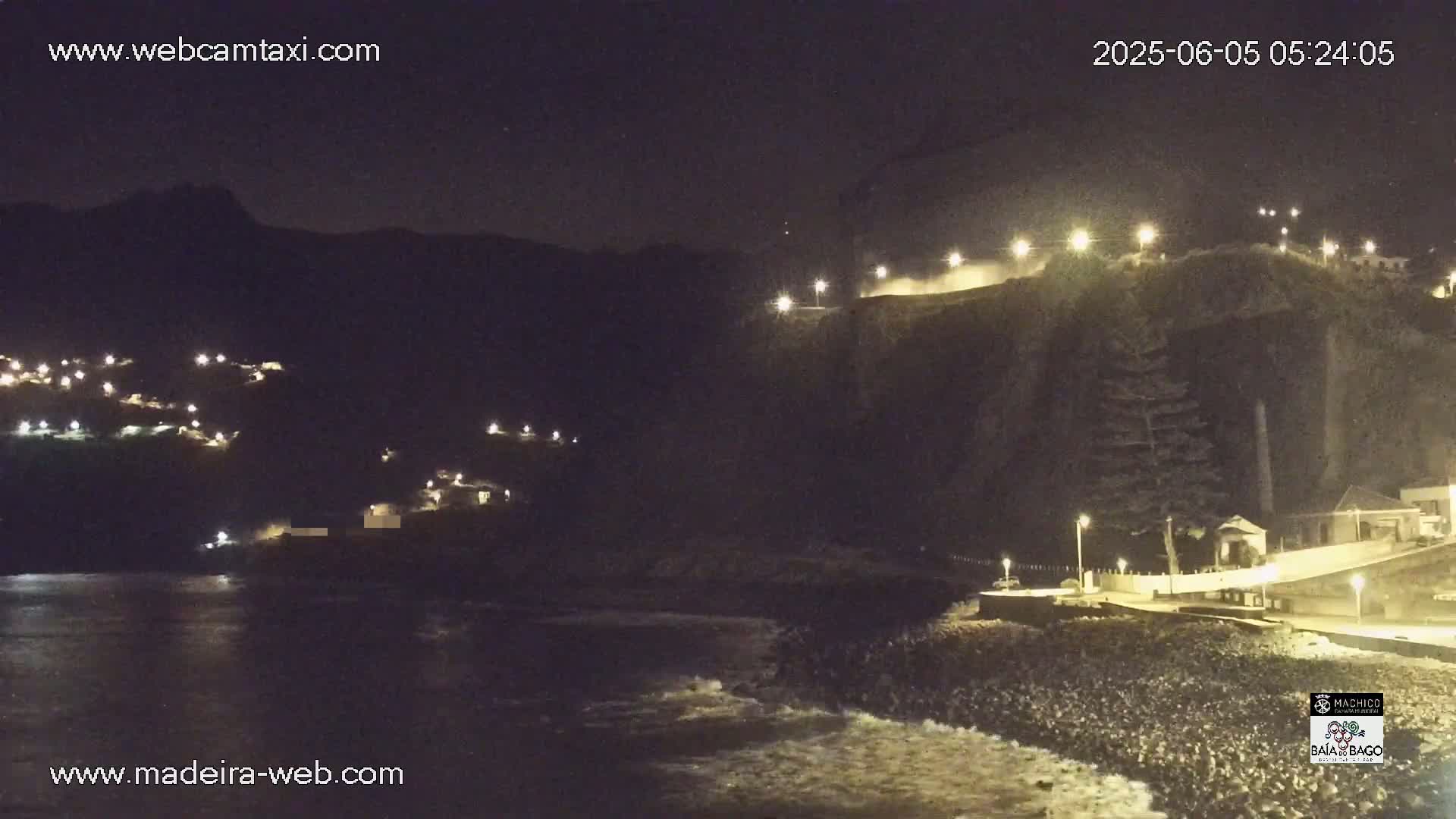 A nighttime coastal scene shows a dark, rocky cliff face illuminated by streetlights, with a small town nestled on the hillside above a dark ocean with small waves.