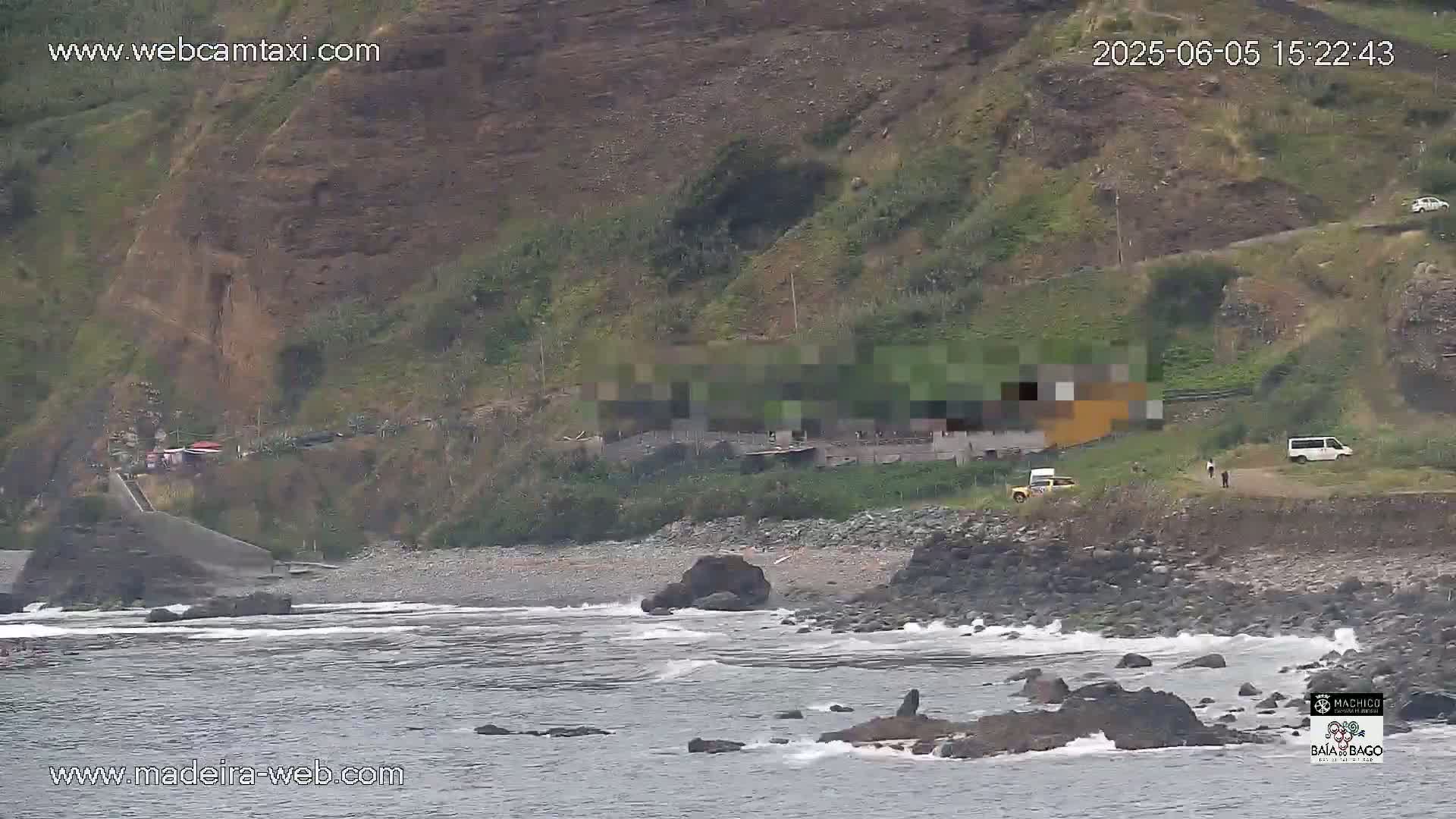 A rocky coastline with a dark-colored beach, some buildings on a hillside, and a few vehicles parked near the water's edge under overcast skies.