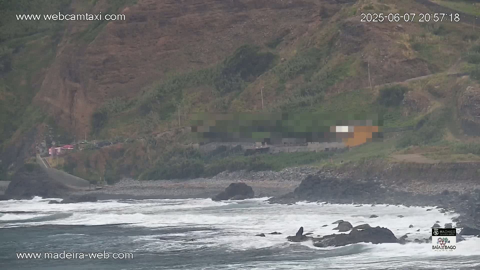 Waves crash against a rocky shoreline below a steep, green hillside with a few buildings near the top.