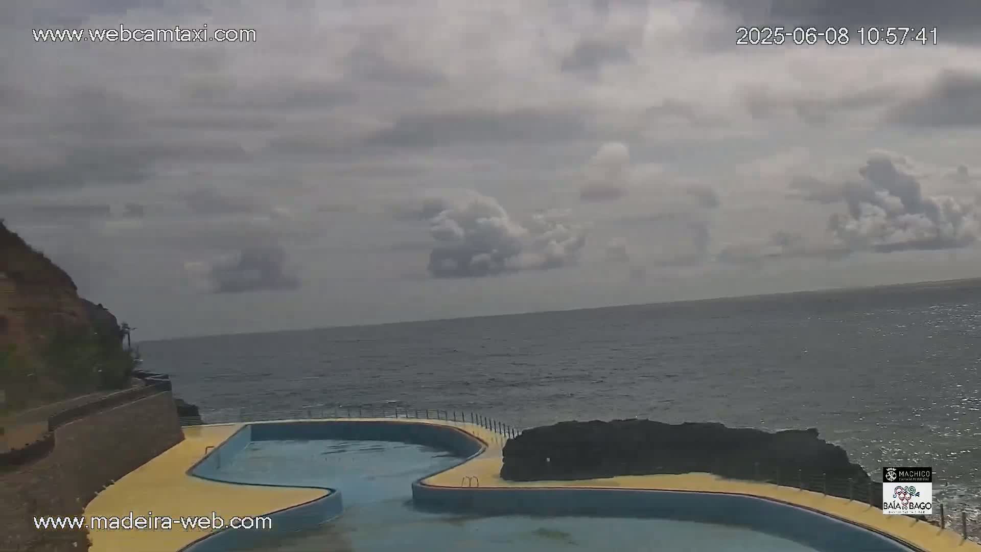 An outdoor scene shows a curved, oceanside pool next to a rocky cliff under a mostly cloudy sky.