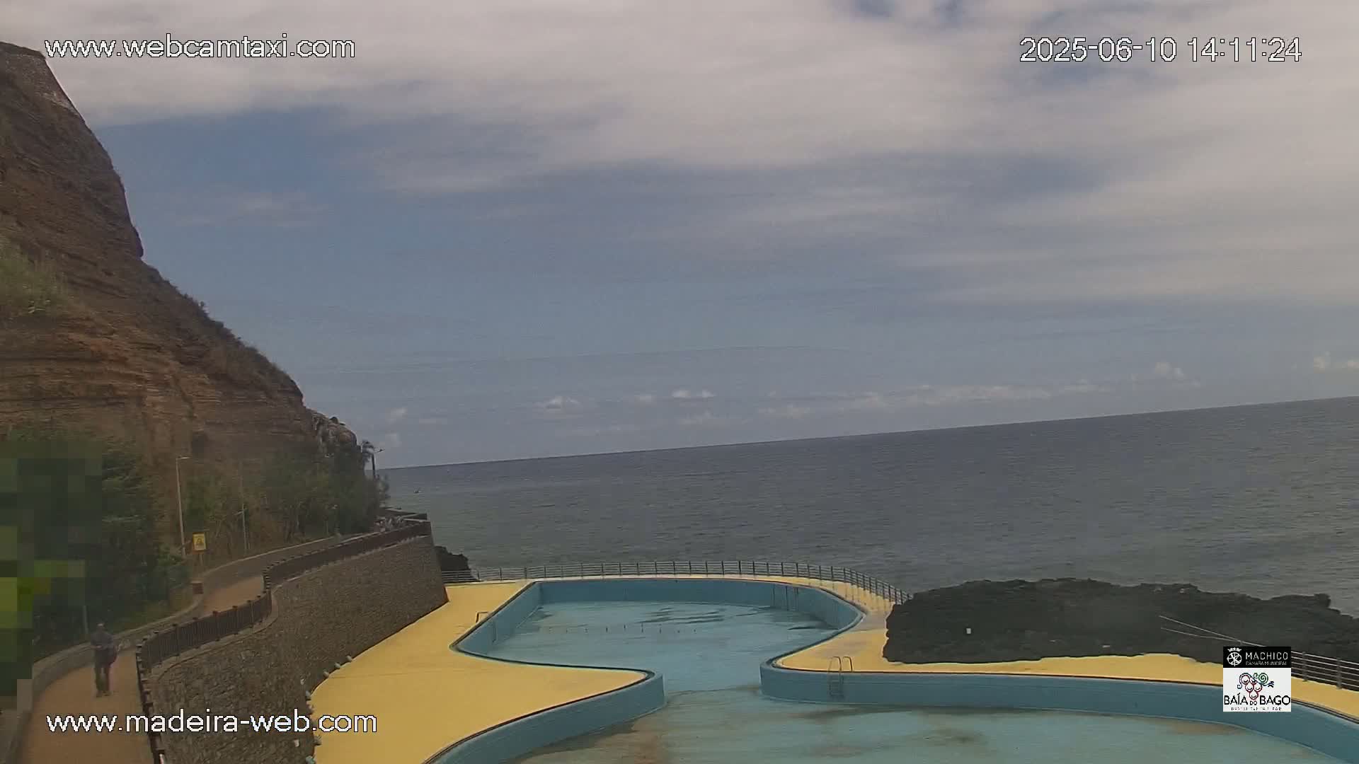 An outdoor view shows a partly cloudy sky over a calm ocean, with a curved, empty swimming pool next to a rocky cliff and a paved walkway.