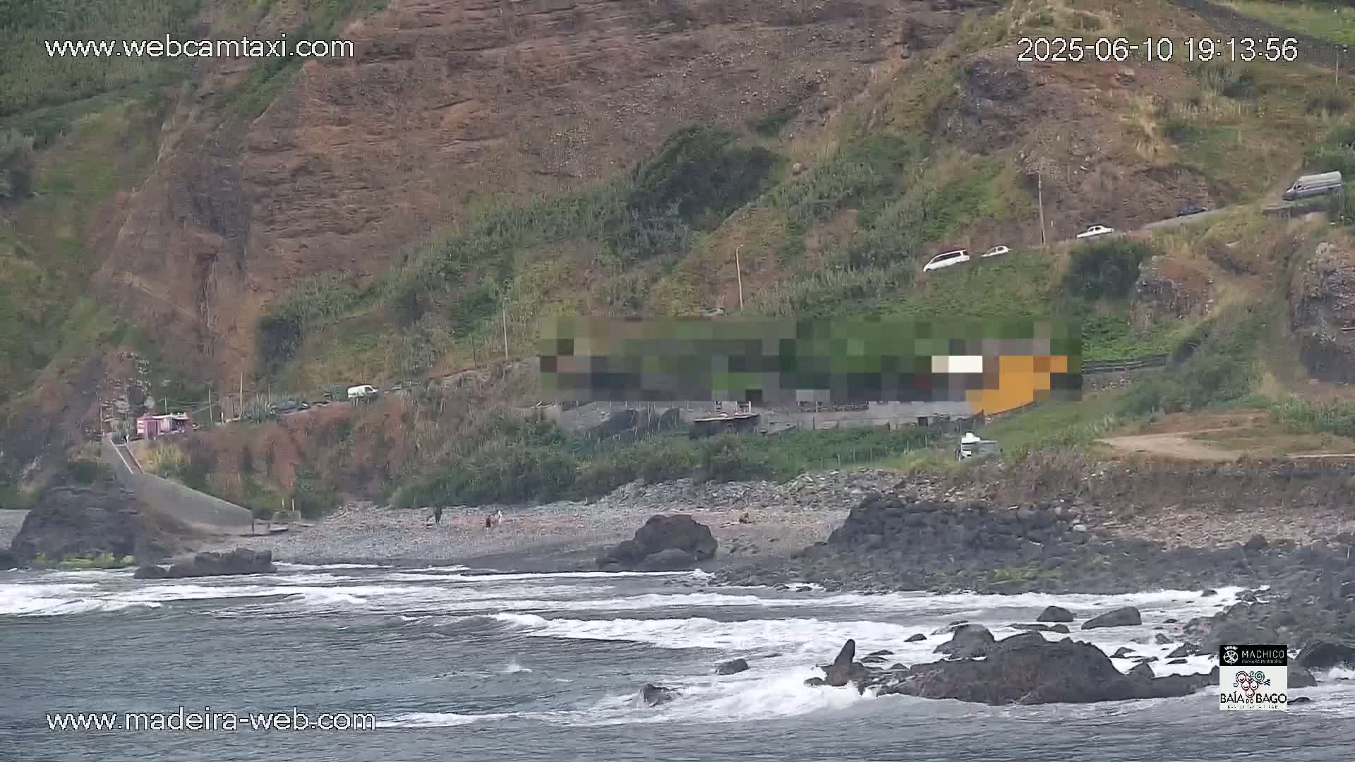 A rocky beach with dark sand and small waves laps against the shore, under an overcast sky, backed by a steep, green hill with a few cars parked along a road at its crest.