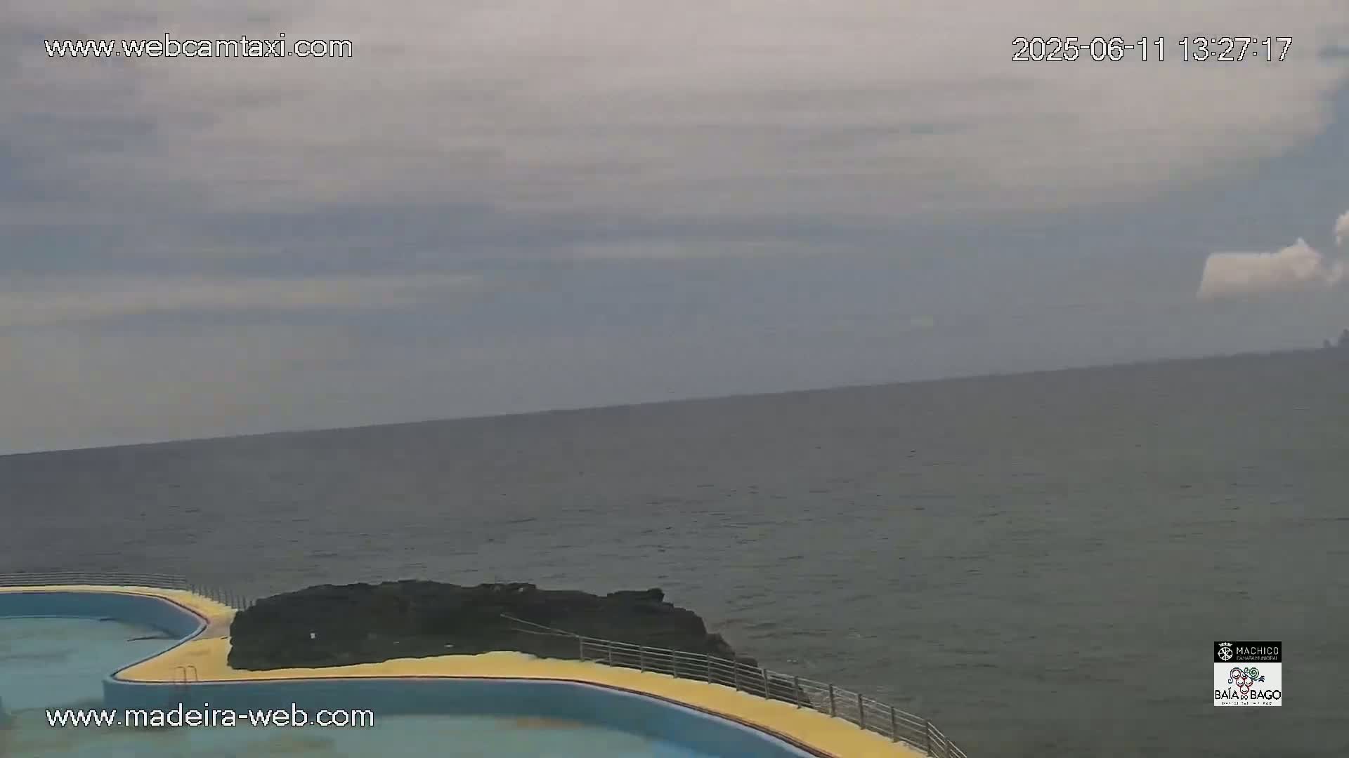 An outdoor view of a calm ocean under a cloudy sky, partially encompassing a yellow-edged pool adjacent to dark rocks.