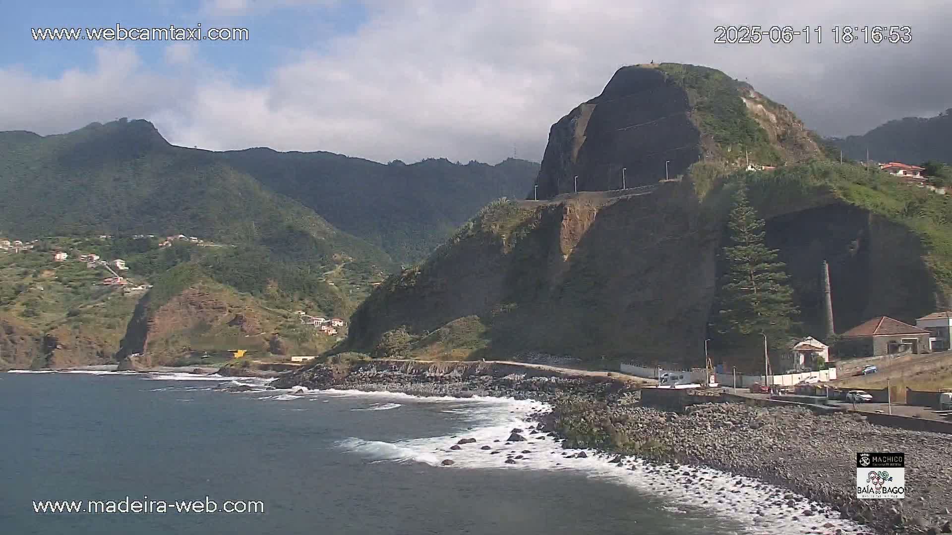 A rocky coastline with a large cliff face and a small town nestled against a backdrop of mountainous terrain under partly cloudy skies.