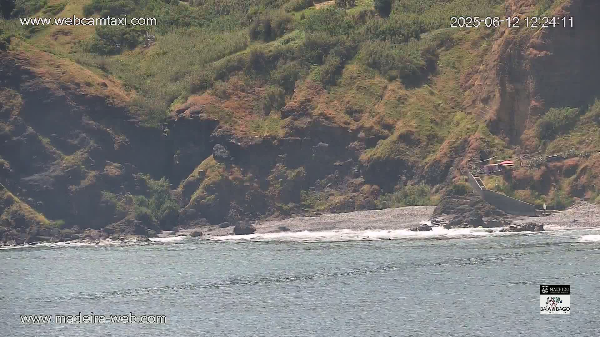 A rocky, vegetated hillside slopes down to a small, pebble beach where small waves lap the shore on a sunny day.