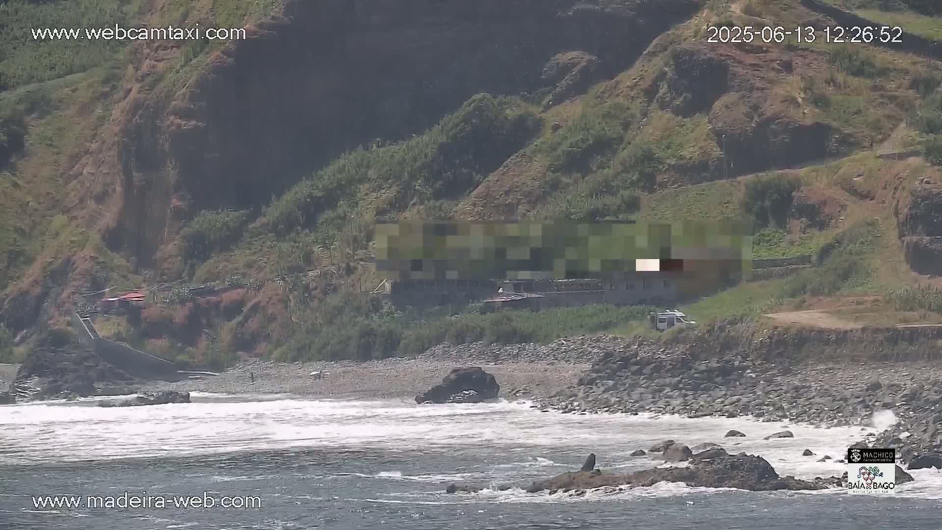 A rocky shoreline with a small beach and a building on a hillside is seen under sunny conditions, with waves breaking in the ocean.