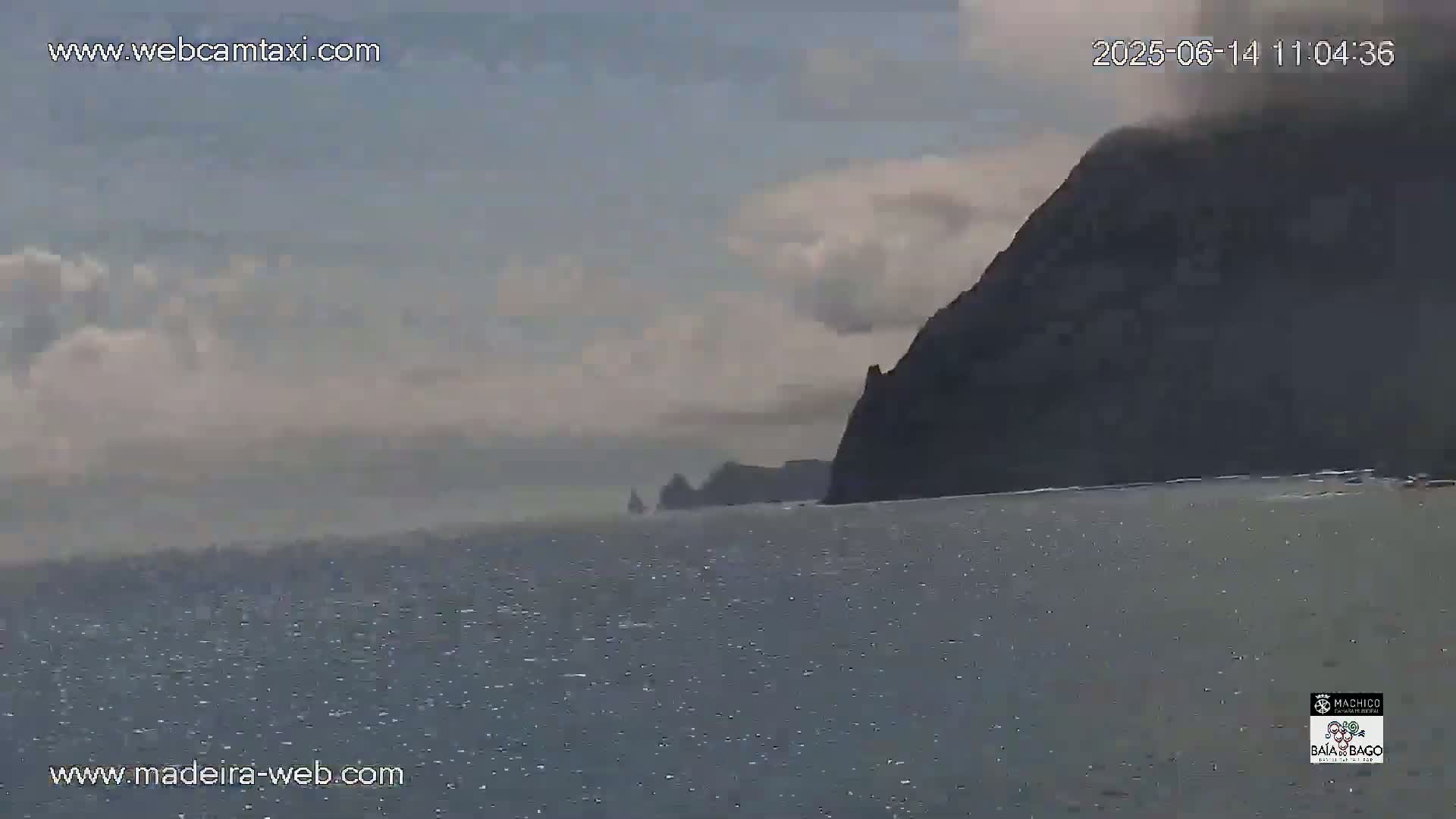A dark, rocky cliff face rises from a calm, gray ocean under a cloudy sky.