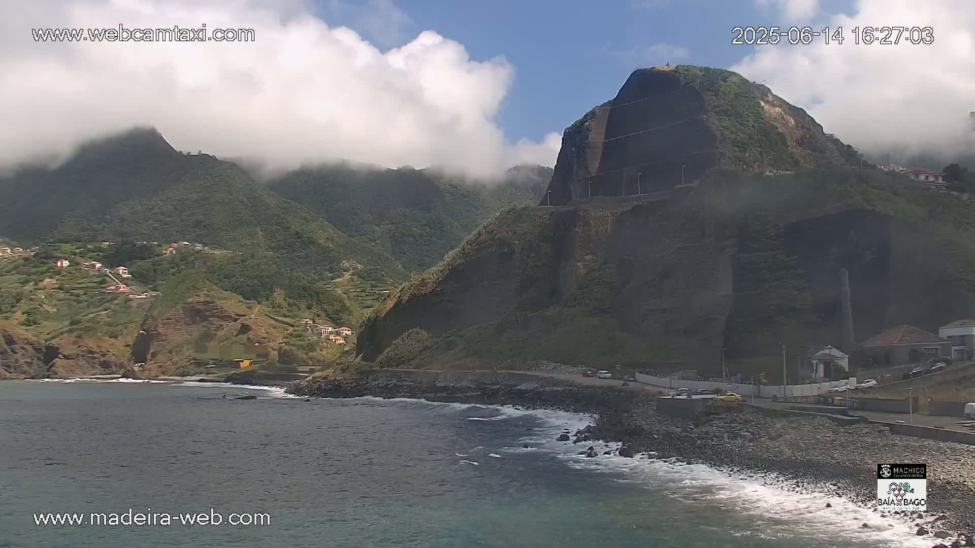 A partly cloudy day reveals a rocky coastline with a dark, steep cliff face overlooking a calm sea, and a village nestled into the green hills behind it.