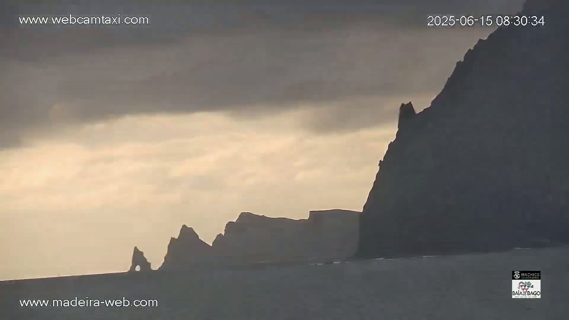 Silhouetted cliffs rise from a calm sea under a cloudy, pale sky.